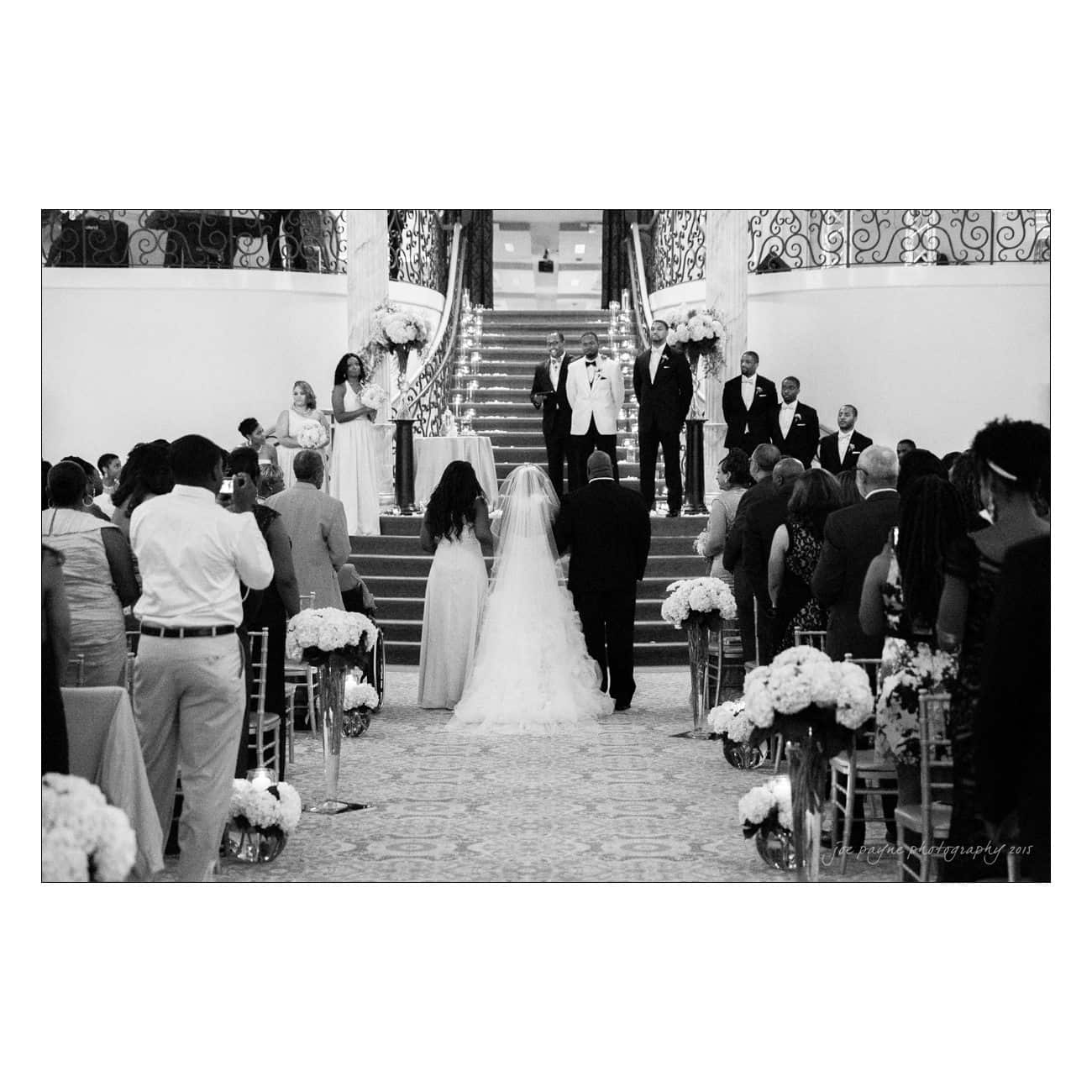 A black and white photo captures a wedding ceremony. The bride and groom stand at the altar, surrounded by the wedding party and guests. Floral arrangements line the aisle, leading to a grand staircase in the background.