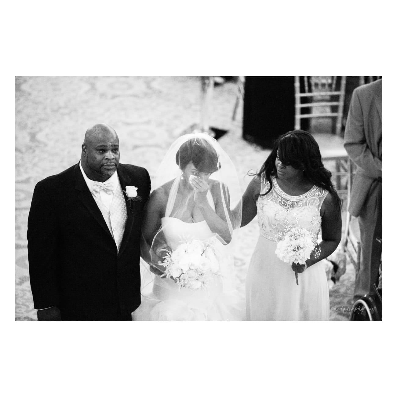 A black and white image of a bride in a wedding dress and veil walking down the aisle, accompanied by two people, one on each side. The bride holds a bouquet and appears emotional. The scene is captured from a slightly elevated angle.