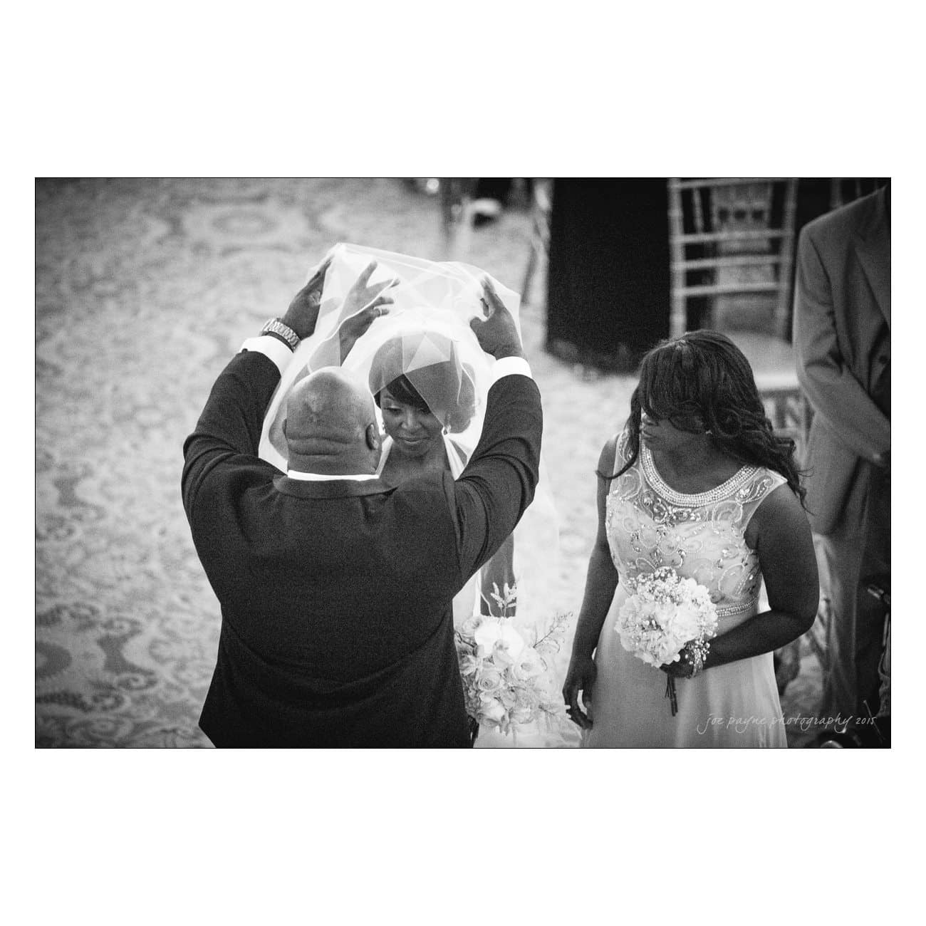 A black and white photo of a man lifting a brides veil during a wedding ceremony. The bride holds a bouquet, and a woman stands beside her, also holding flowers. Elegant chairs are visible in the background.
