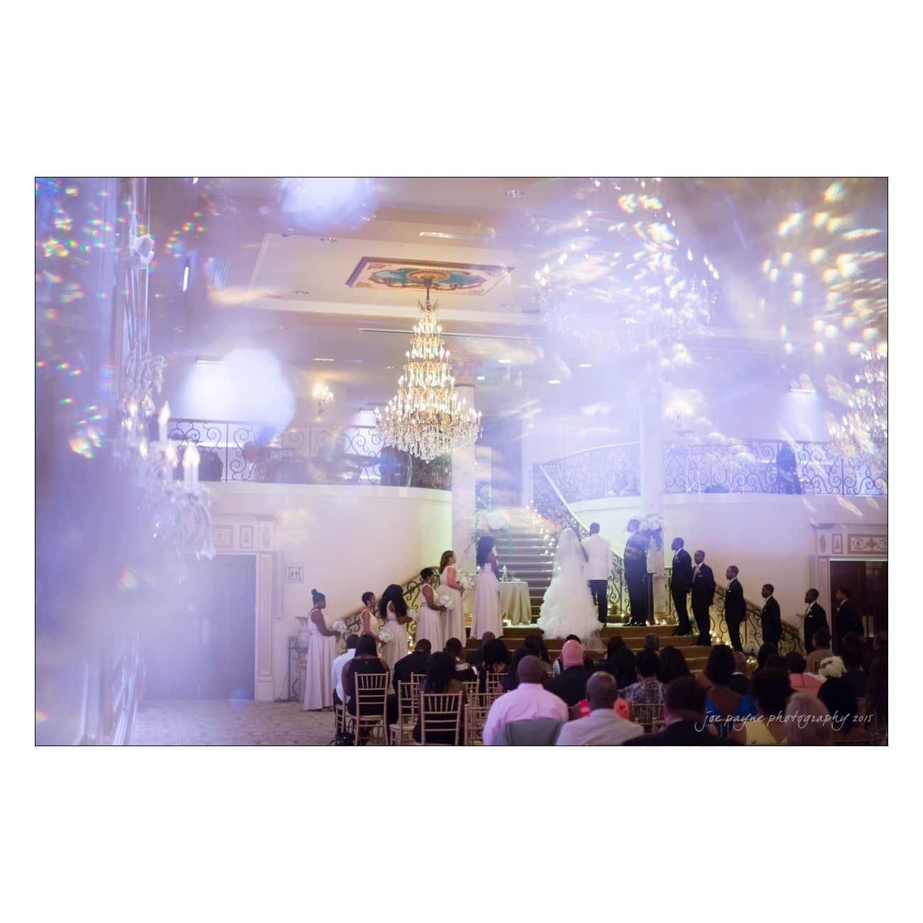 A wedding ceremony in a grand indoor venue, featuring a couple on a staircase surrounded by their wedding party. Guests sit nearby, and a large chandelier hangs from the ceiling, with colorful lights creating a dreamy atmosphere.