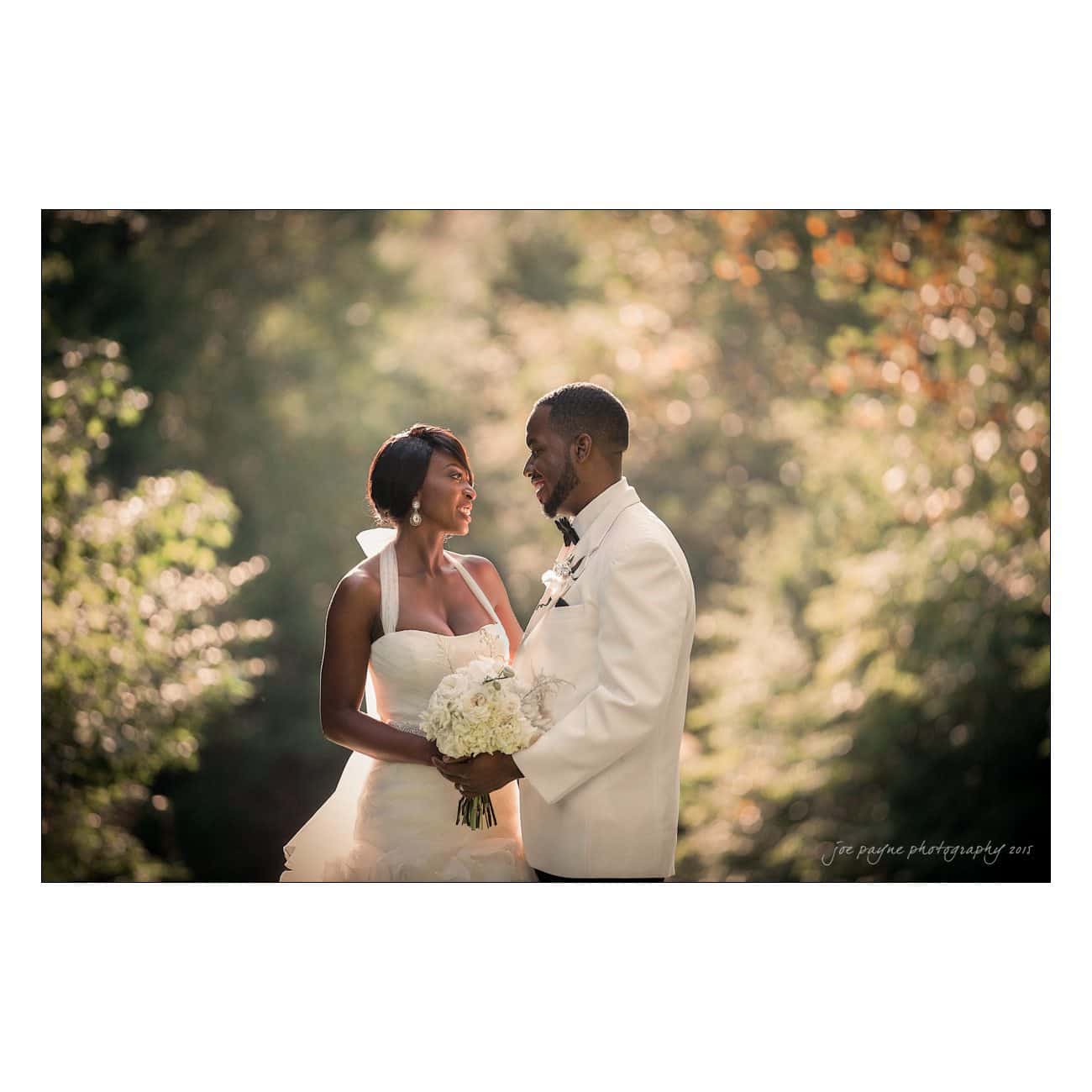 A couple is dressed in wedding attire, smiling at each other. The bride holds a bouquet of white flowers. They are outdoors with greenery and soft, natural lighting in the background, creating a romantic atmosphere.
