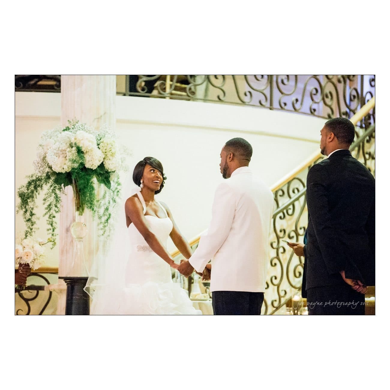 A bride and groom hold hands and gaze at each other during their wedding ceremony, with a groomsman standing nearby. The setting includes elegant floral arrangements and an ornate staircase in the background.