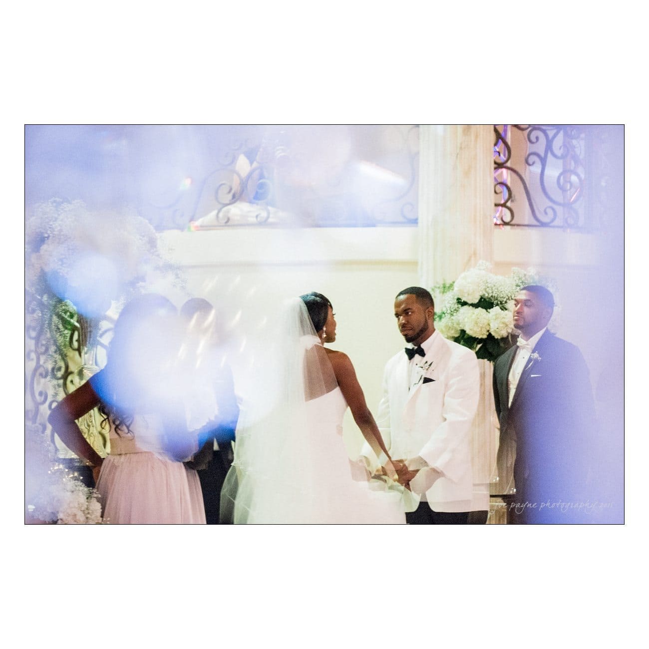 A bride and groom stand facing each other during their wedding ceremony, surrounded by bridesmaids and groomsmen. The scene is softened by a dreamy, blurred effect, and they are framed by elegant floral arrangements and ornate decor.