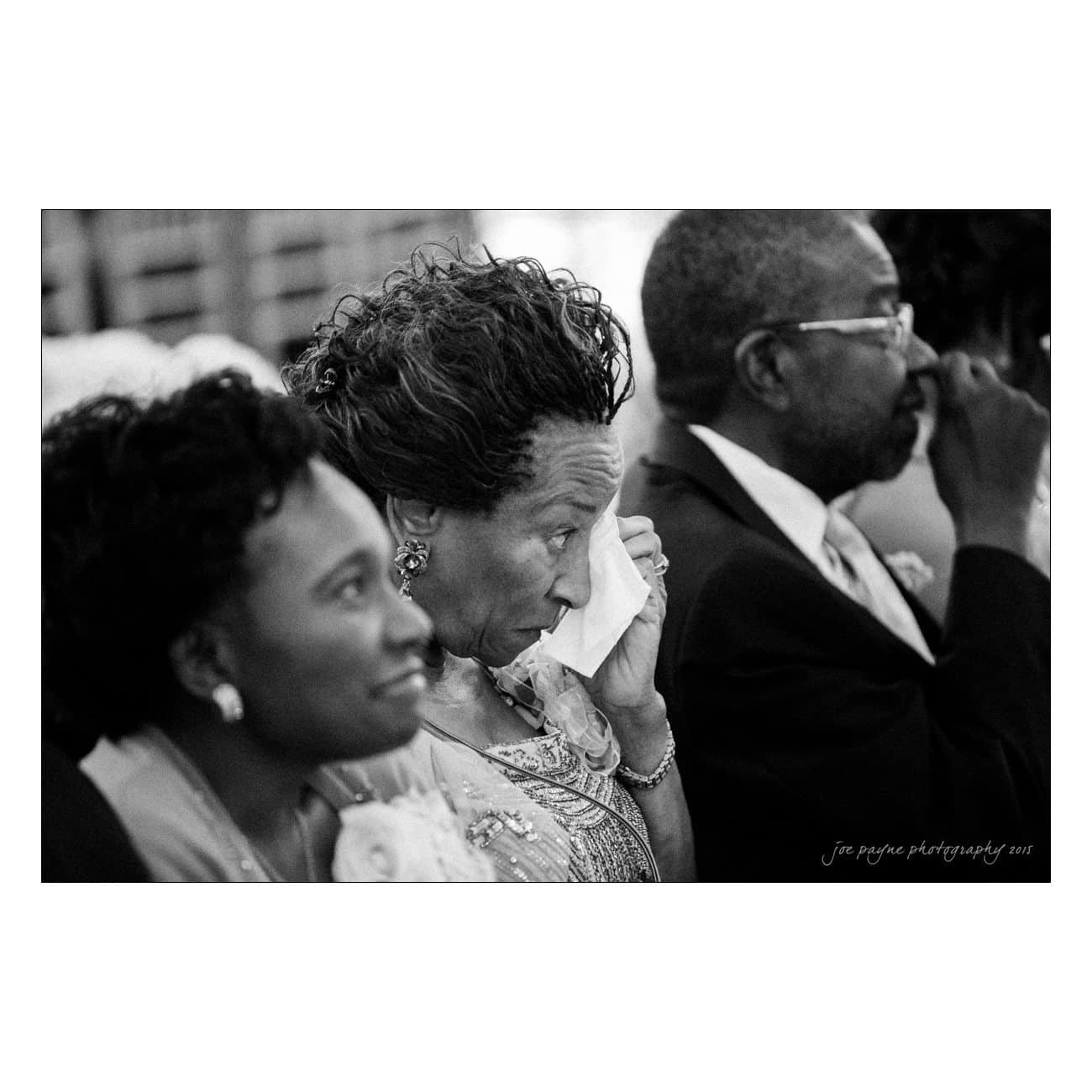A group of three people sits in a row, dressed in formal attire. The woman in the middle dabs her face with a tissue, looking emotional. The setting appears to be a special occasion or ceremony.