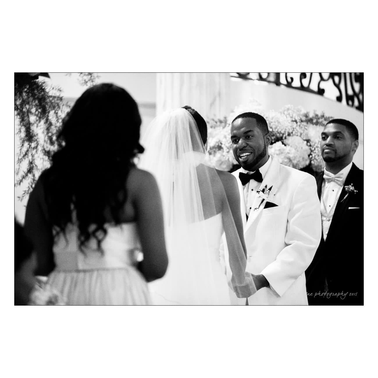 A groom in a white suit smiles at his bride, who is wearing a veil and a strapless dress, as they stand at an altar. Another man and a woman in formal attire are present, surrounded by floral decorations.