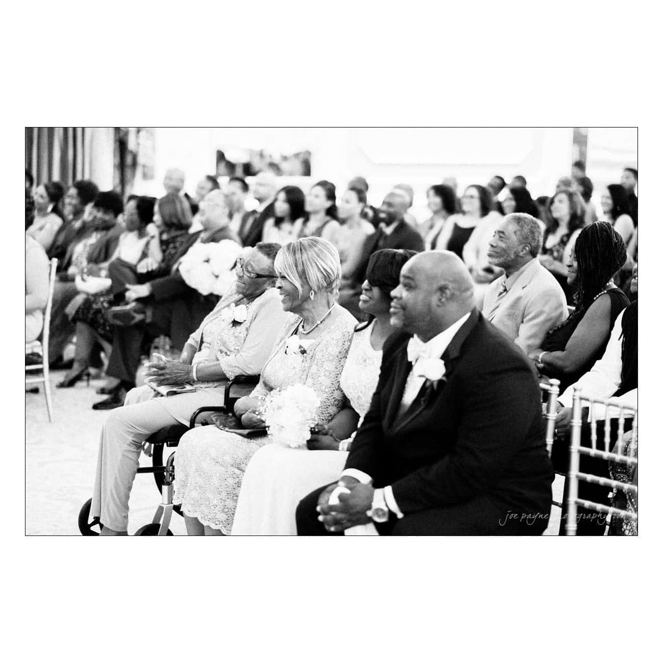A black and white photo of an audience seated at an event. Front row has smiling attendees, including a man in a suit and women holding flowers. The setting appears formal, with many people in the background.
