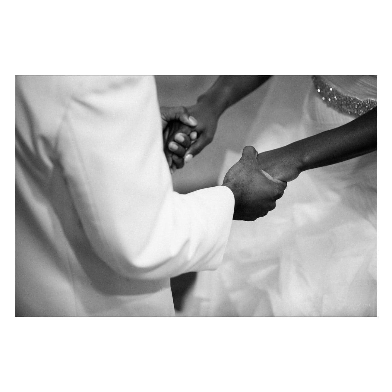 A black and white photo showing a couple in formal attire holding hands. The person on the left wears a light suit, while the person on the right wears a dress with a beaded belt. Their clasped hands are the focal point.