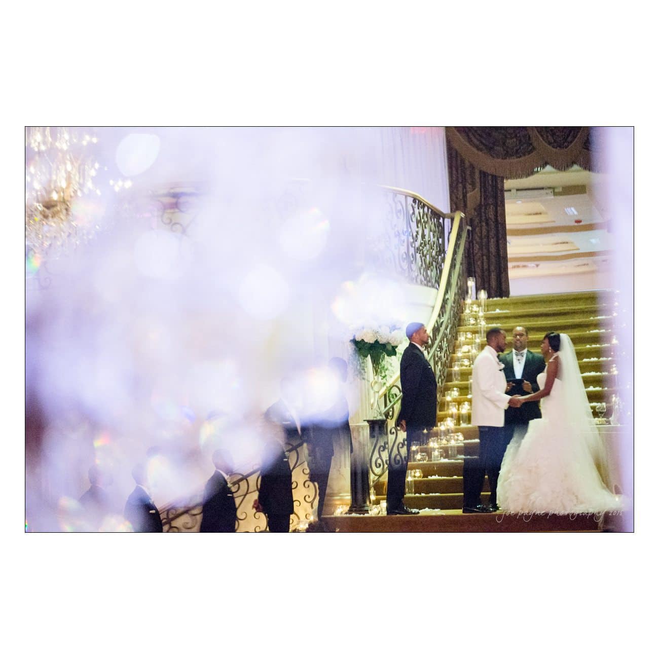 A bride and groom stand at the altar during a wedding ceremony, facing each other while holding hands. The background features a grand staircase with decorative lighting. A priest and two men are present. The forefront is blurred with sparkling lights.