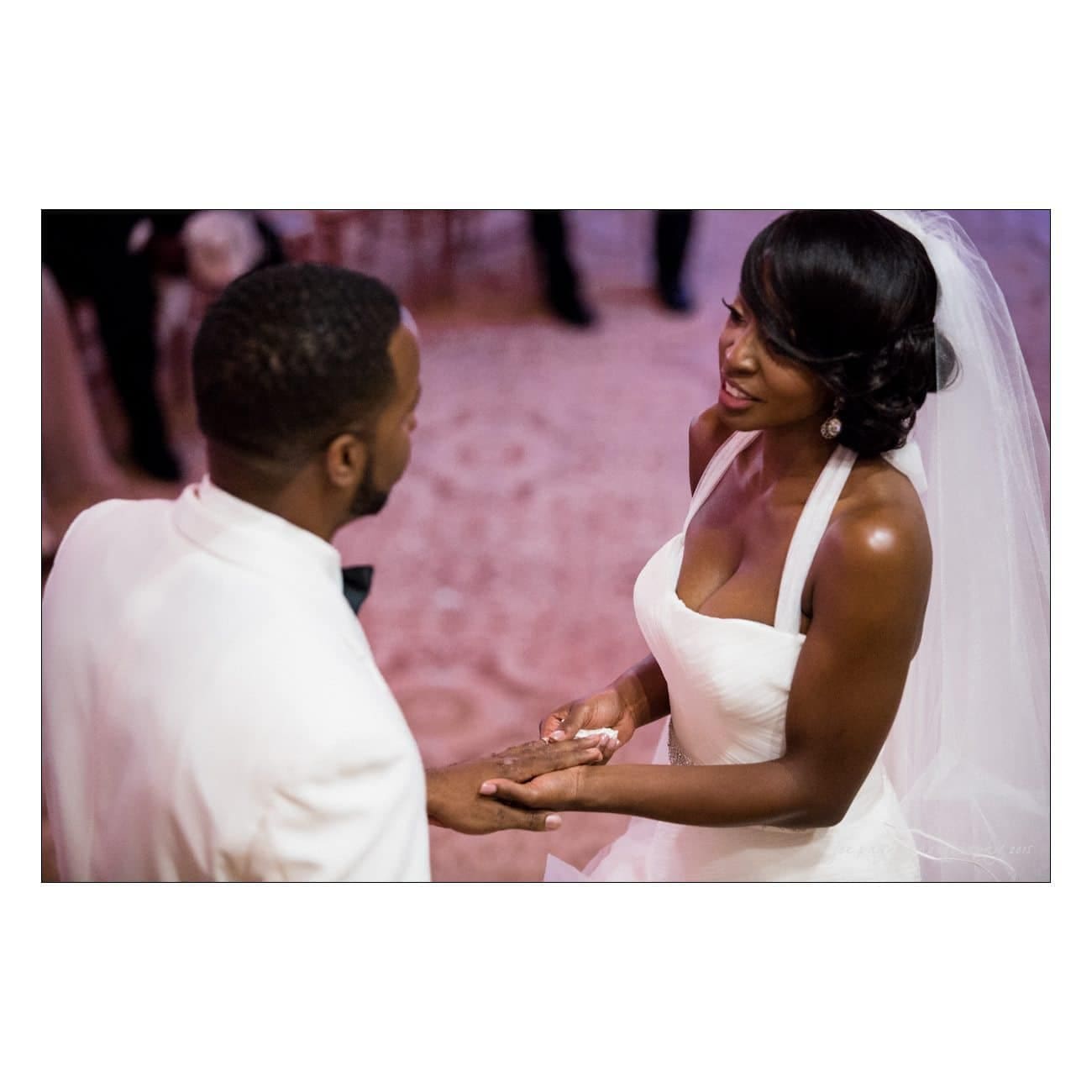 A bride and groom exchange rings during a wedding ceremony. The bride is wearing a white dress with a veil, while the groom is in a white suit. They are looking at each other affectionately.