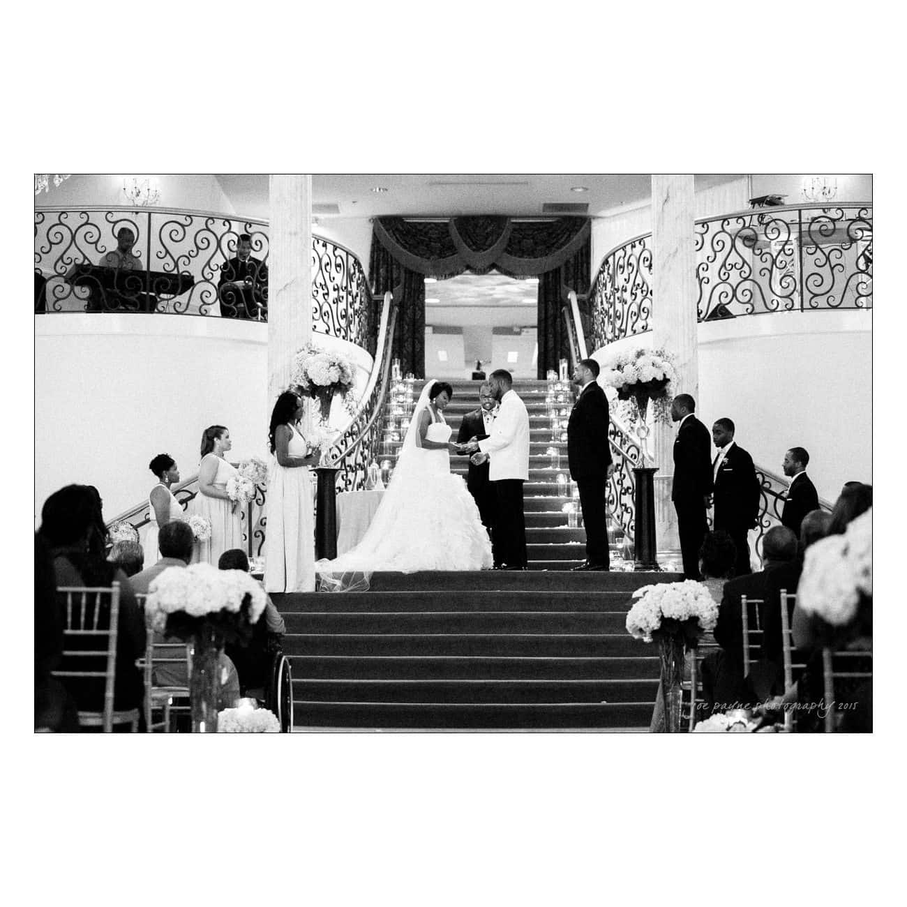 A black and white photo of a wedding ceremony at the bottom of a grand staircase. The bride and groom stand in the center flanked by bridesmaids and groomsmen, with guests seated on either side. Large floral arrangements decorate the scene.