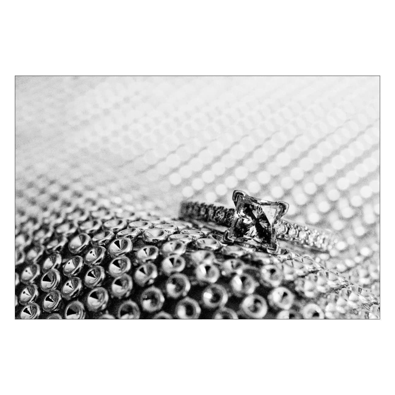 Black and white close-up of a diamond ring with a square-cut stone. The ring rests on a shimmering textured surface with a pattern of small circles, creating an elegant and sophisticated look.