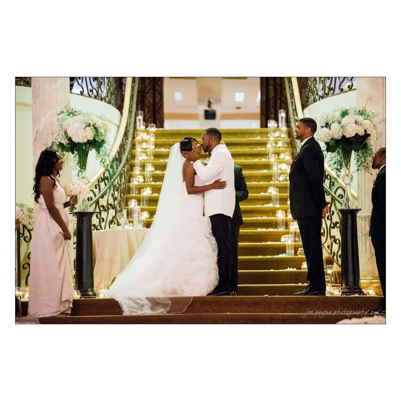 A couple shares a wedding kiss at the base of a grand staircase adorned with white flowers and candles. The bride wears a flowing white dress and the groom is in a white jacket. Bridesmaid and groomsmen stand on either side, watching the ceremony.