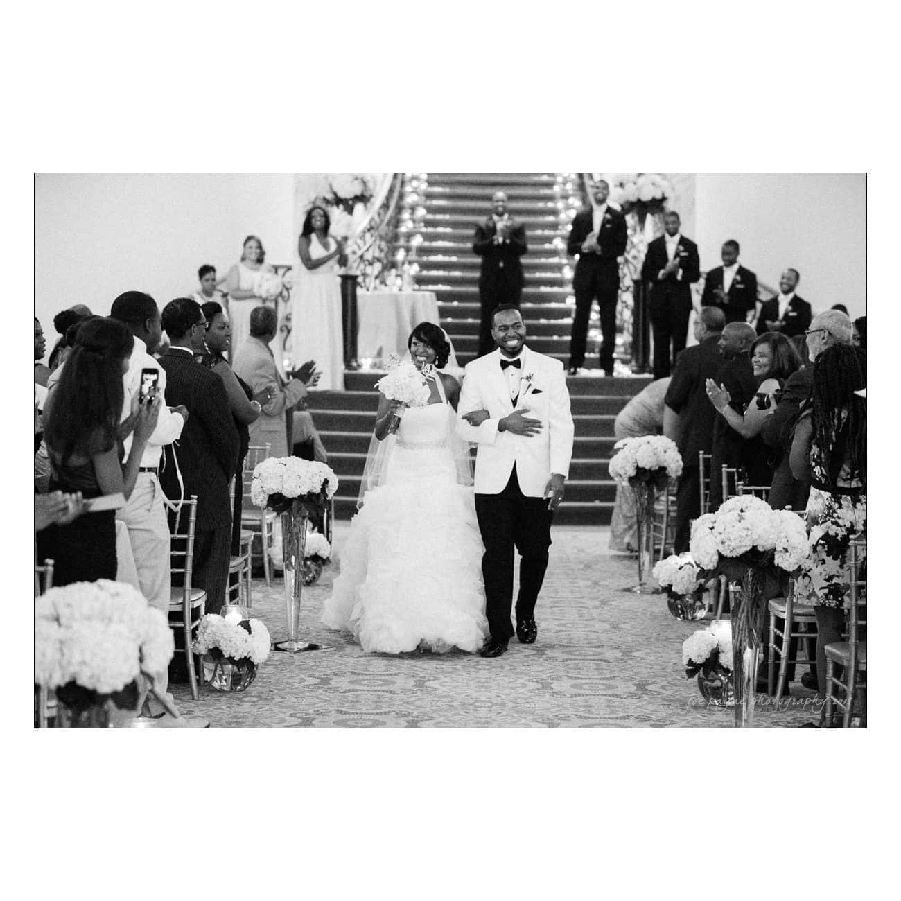 A bride and groom joyfully walk down the aisle after their wedding ceremony, surrounded by applauding guests. The setting is elegant, with floral arrangements and a staircase adorned with lights in the background.