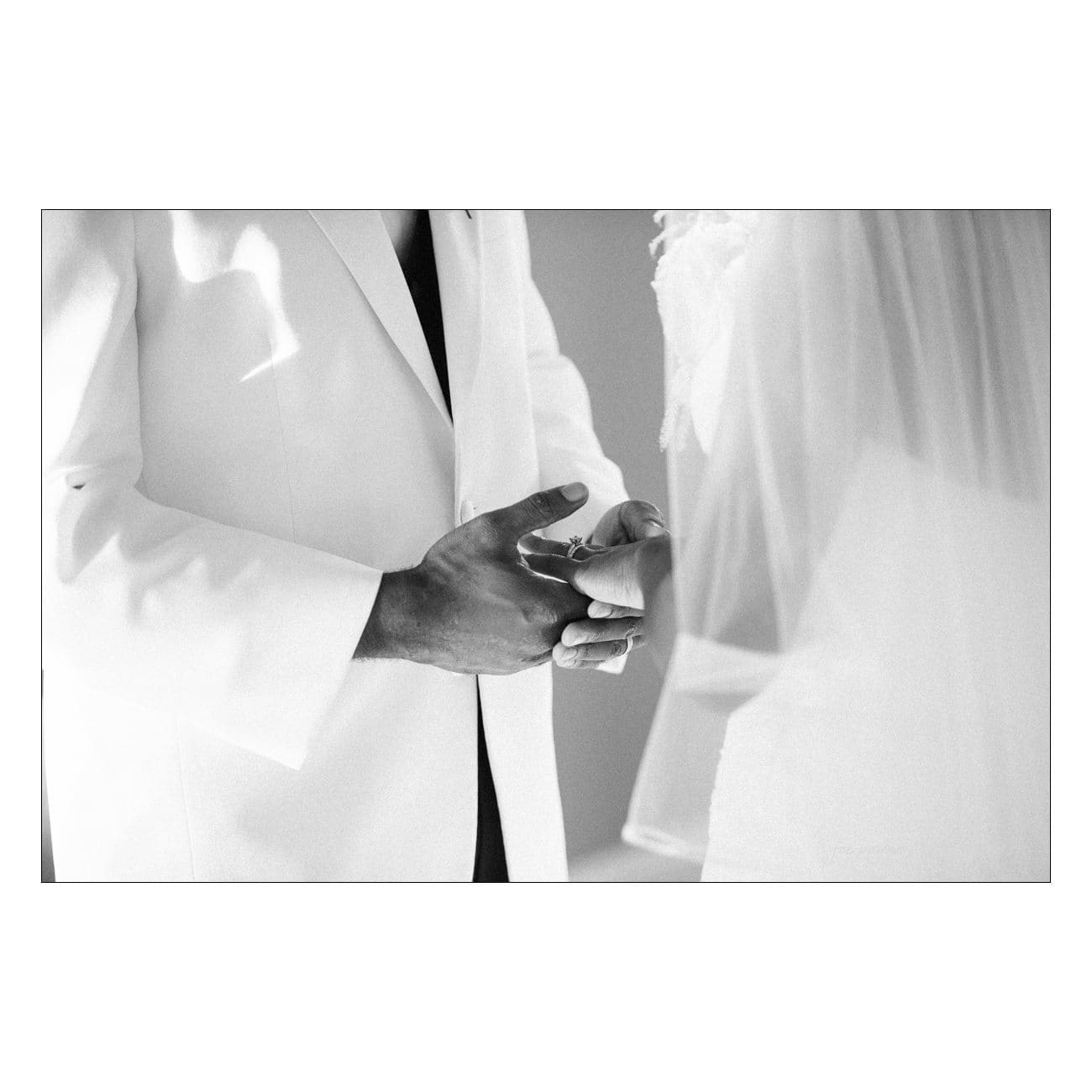 A black and white photo of two people exchanging rings. One person wears a light-colored suit jacket, and the other is dressed in a veil. The focus is on their hands as the ring is being placed.