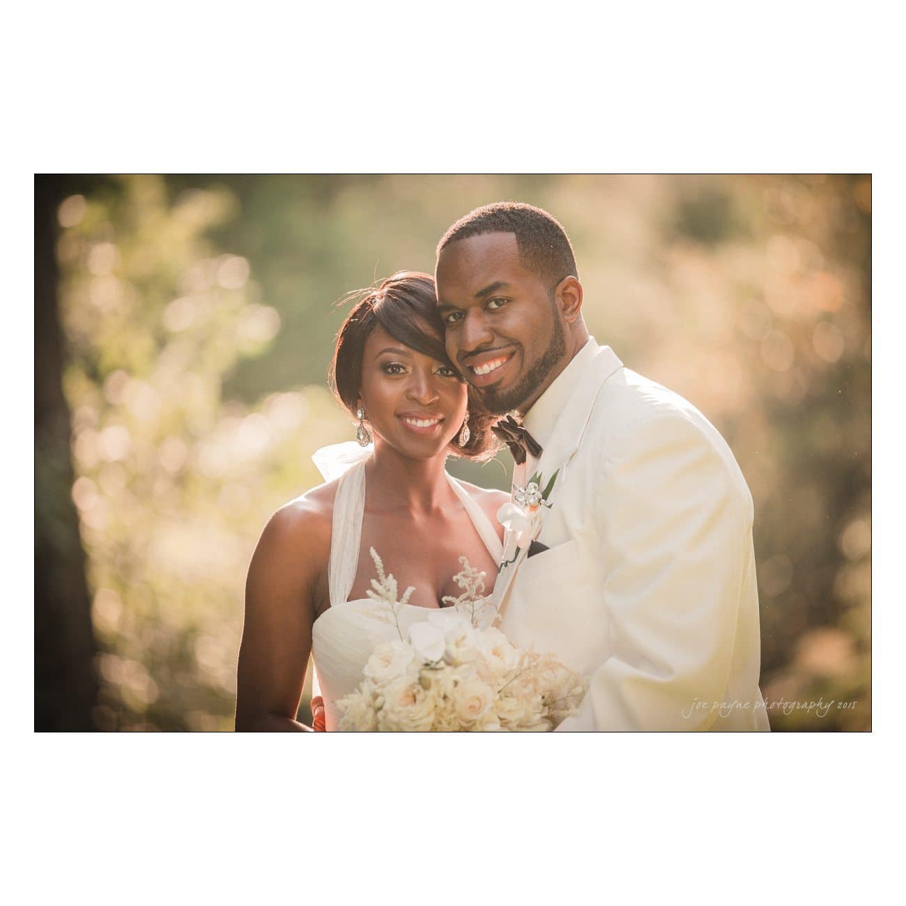 A bride and groom smile warmly at the camera. The bride holds a bouquet of white roses, wearing a white gown with a halter neckline. The groom is in a white suit with a black bow tie. They stand in front of a softly blurred, sunlit background.
