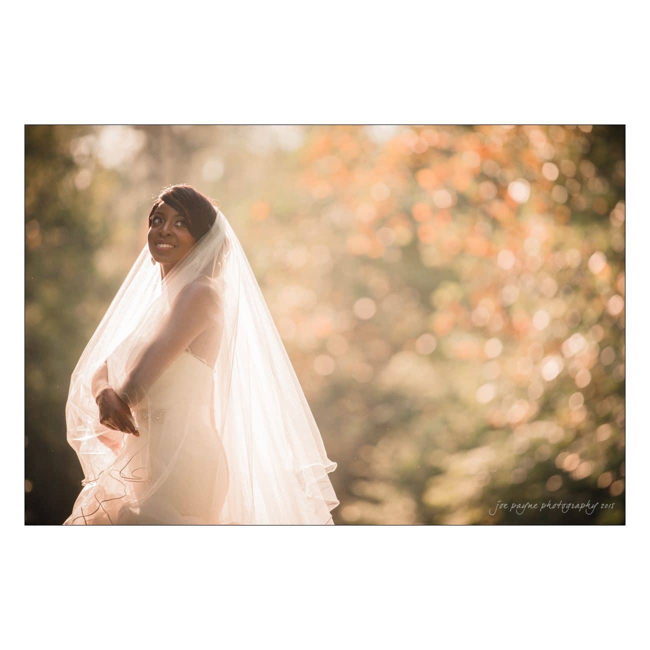 A bride wearing a flowing white veil and a strapless wedding dress stands against a blurred backdrop of trees and sunlight. She is smiling and looking upward, with a serene and joyful expression.