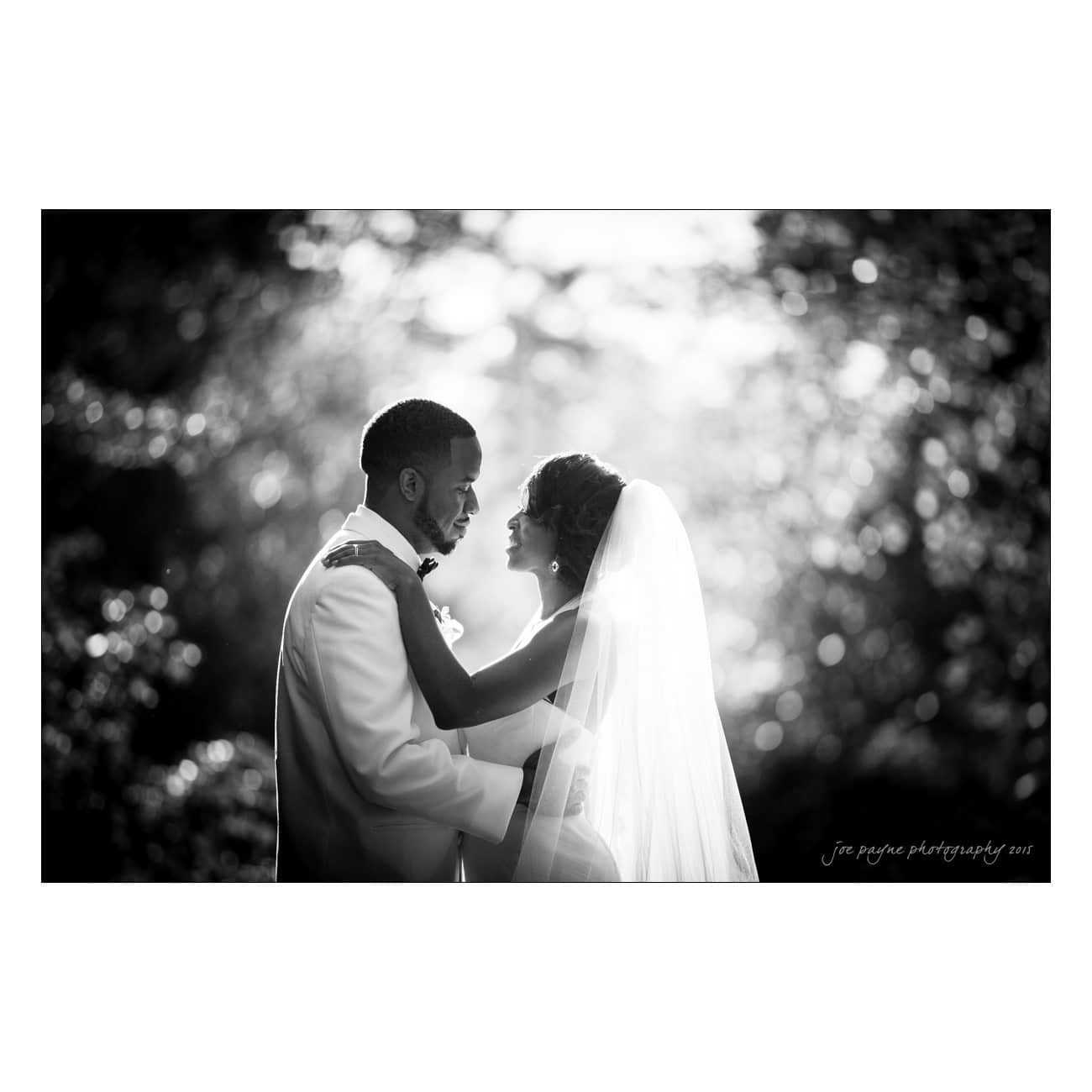 A black and white photo of a newlywed couple embracing outdoors. The groom is in a white suit, and the bride wears a veil. Sunlight filters through the trees, creating a soft, romantic glow around them.