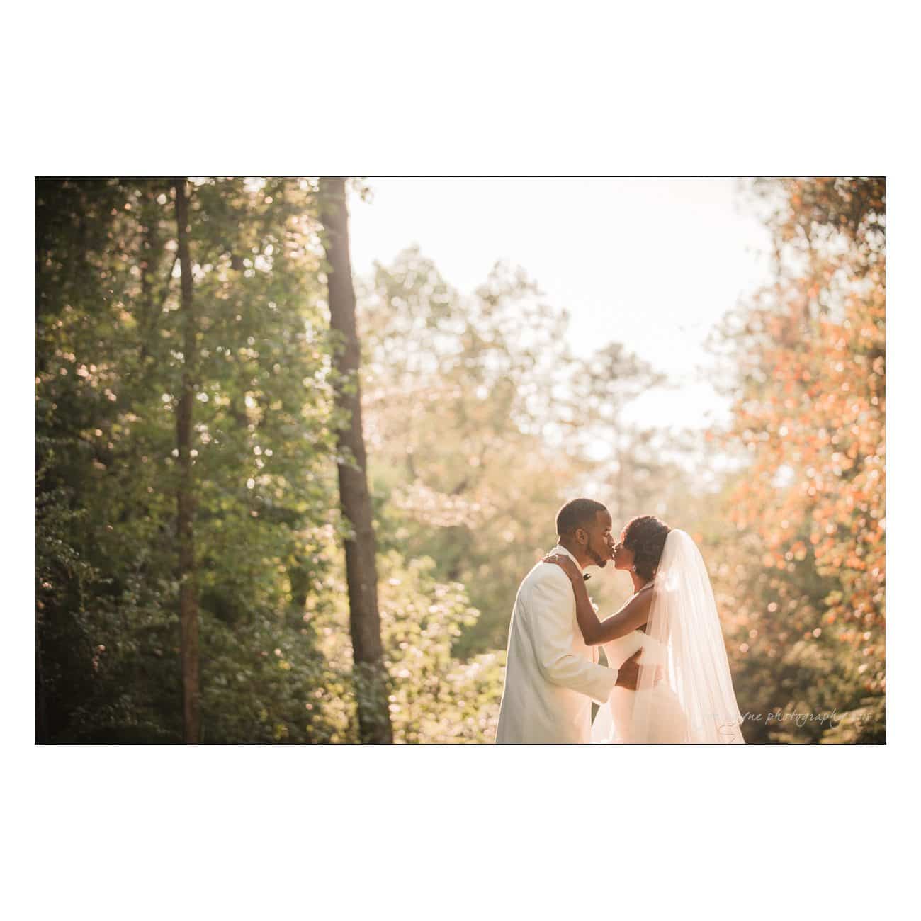 A bride and groom share a kiss in a sunlit forest setting. The bride wears a veil and a white dress, and the groom is in a white suit. Sunlight filters through the trees, creating a romantic atmosphere.