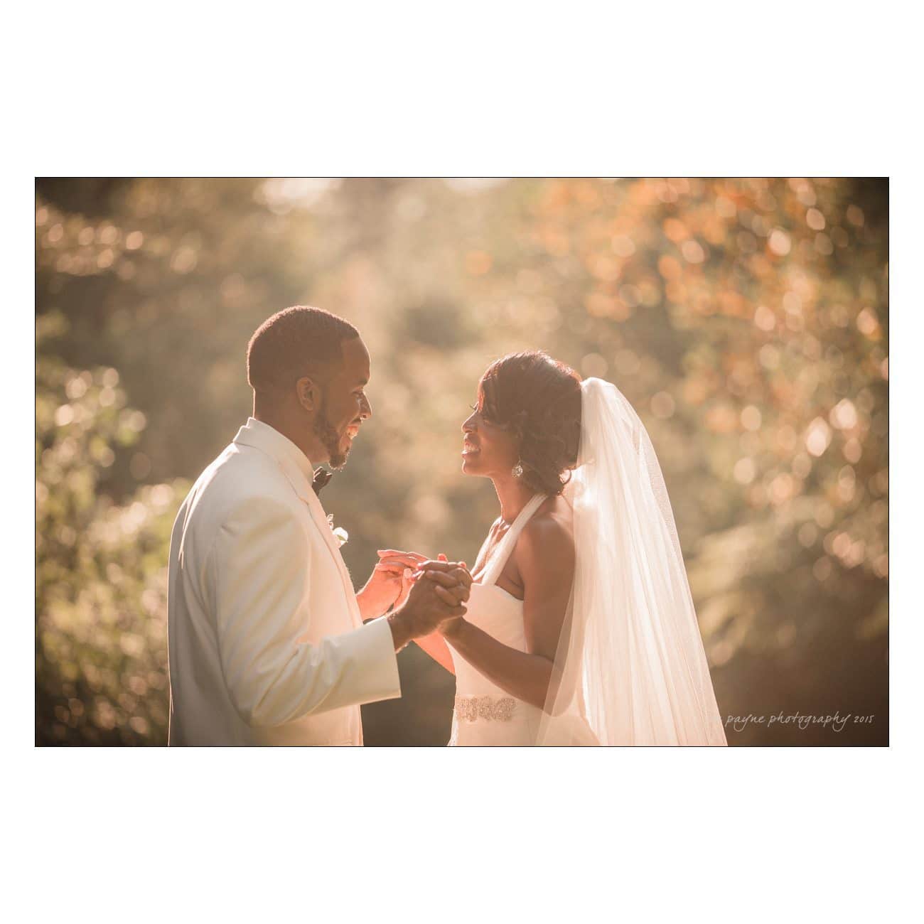 A bride and groom holding hands and smiling at each other outdoors. The bride is wearing a white dress with a veil, and the groom is in a white suit. They are surrounded by soft, natural light and a blurred, leafy background.