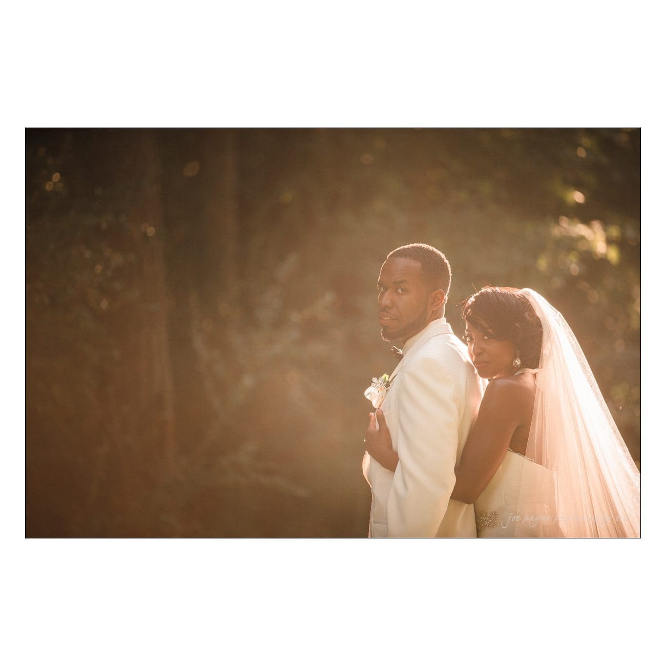 A bride and groom stand close together in an outdoor setting. The bride, in a white dress and veil, rests her head on the grooms shoulder. The groom wears a white tuxedo. Soft, natural light filters through the trees in the background.