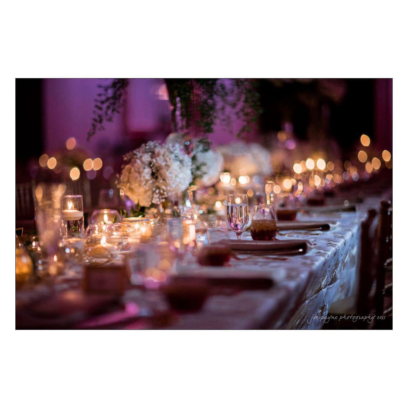 A dimly lit, elegant table setting with white floral centerpieces and glowing candles creating a warm, romantic ambience. Blurred bokeh lights add to the intimate atmosphere of the scene.