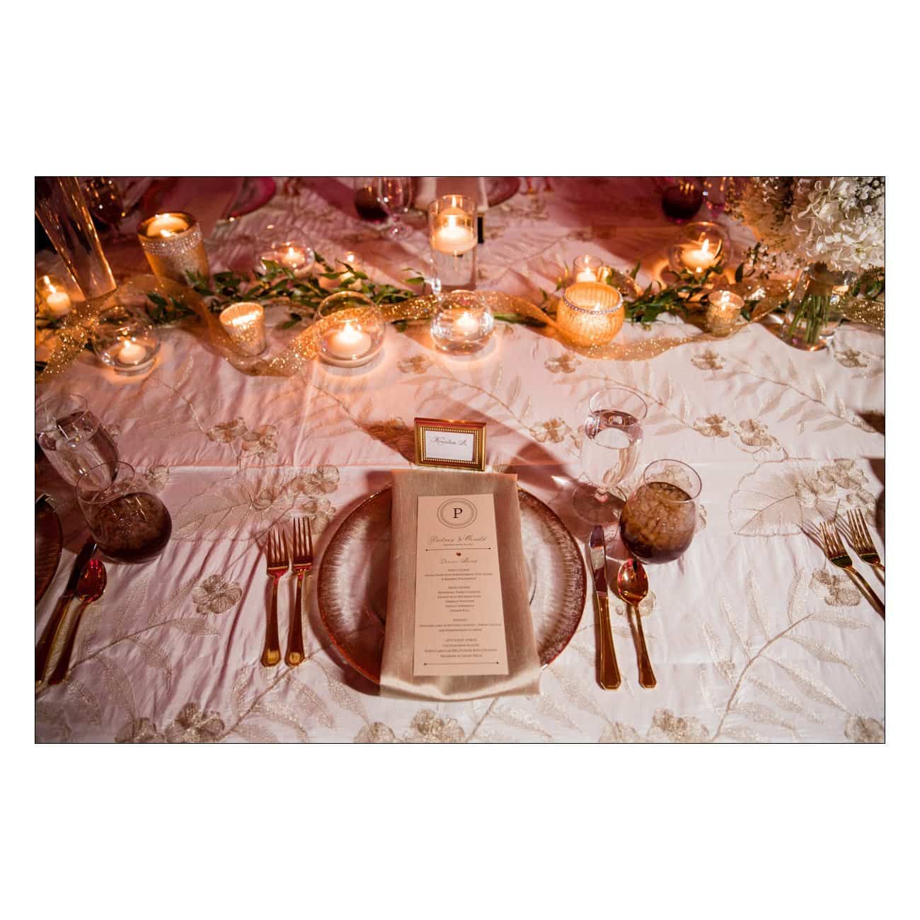 A beautifully set table decorated with candles, greenery, and a white tablecloth with embroidered designs. Theres a place setting with a menu card, gold-colored cutlery, and glassware, creating an elegant and warm atmosphere.