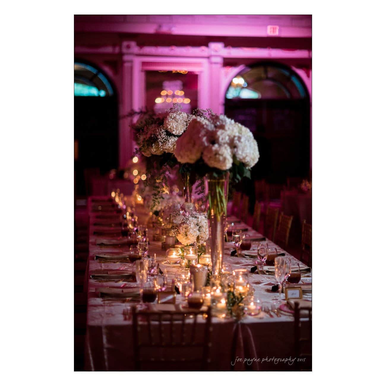 A long, elegantly set dinner table with glowing candles, tall floral centerpieces, and a soft, warm ambiance illuminated by purple lighting in a dimly lit room.
