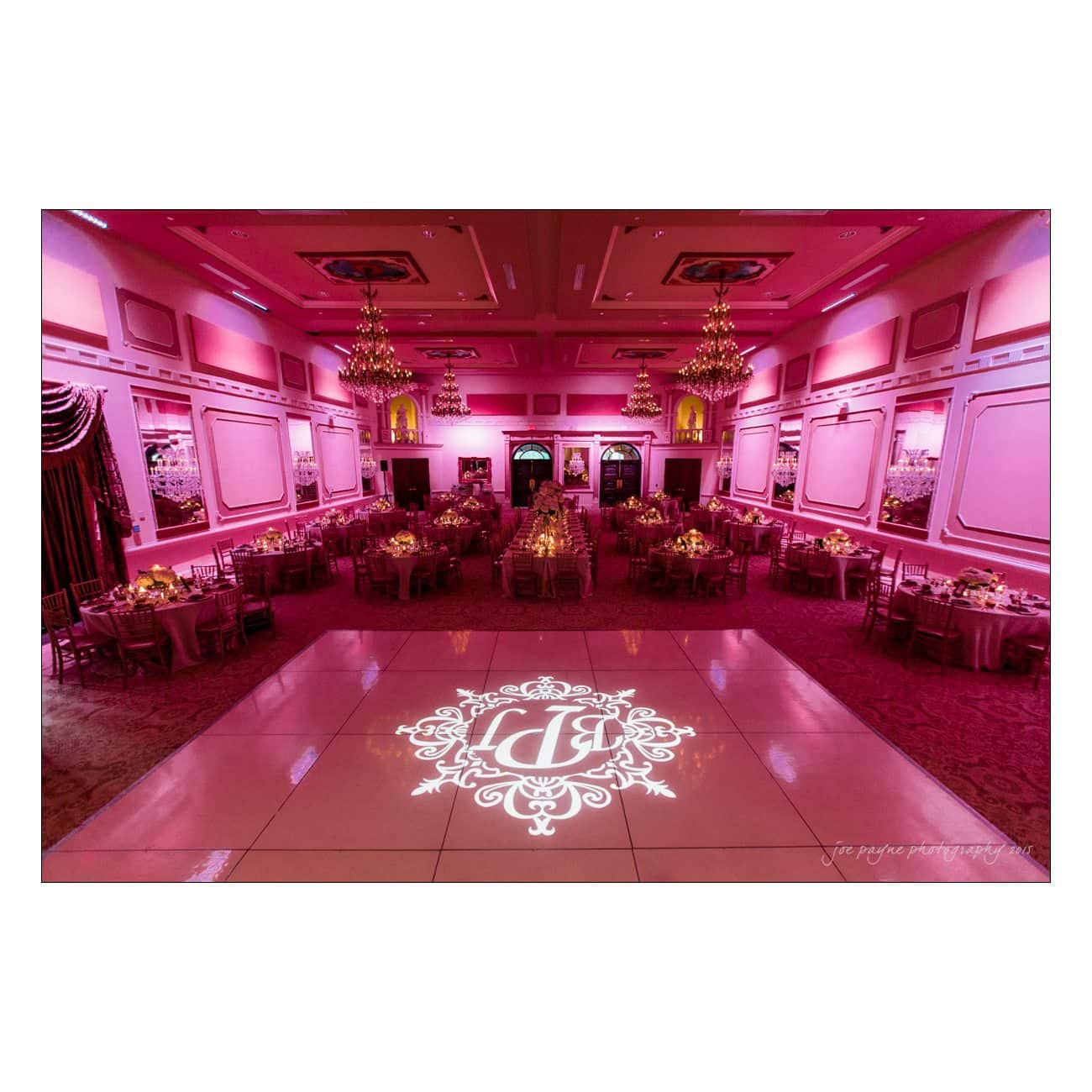 A lavish ballroom set for an event, illuminated in pink lighting. The polished dance floor has an intricate monogram design. Tables are elegantly arranged with floral and candle centerpieces, and the ceiling features ornate chandeliers.