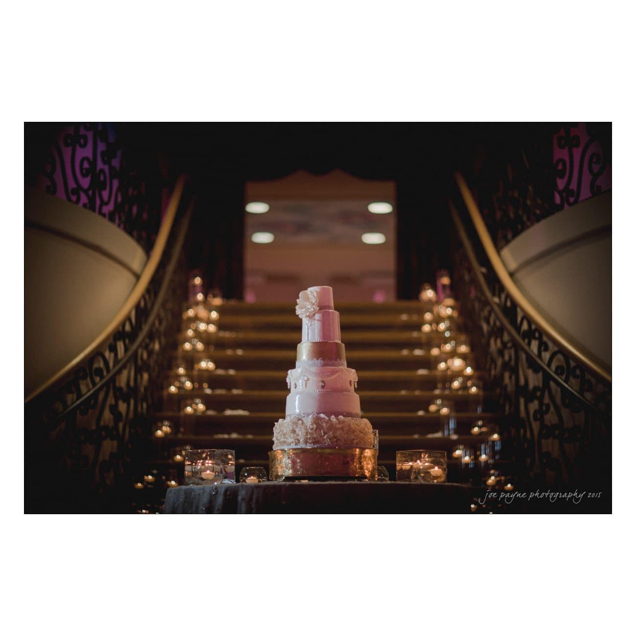 A luxurious, multi-tiered wedding cake with intricate white and pink details stands on a table. It is illuminated by soft lighting, with candlelit decorations lining the elegant, curved staircase behind it.
