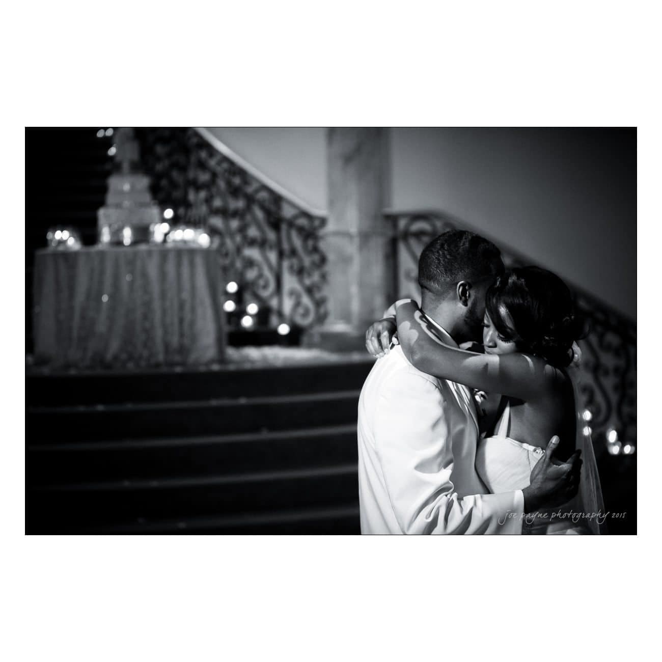 A couple shares a tender embrace while dancing in an elegant setting. The background features a grand staircase and softly glowing lights, adding a romantic ambiance to the scene. The image is in black and white.
