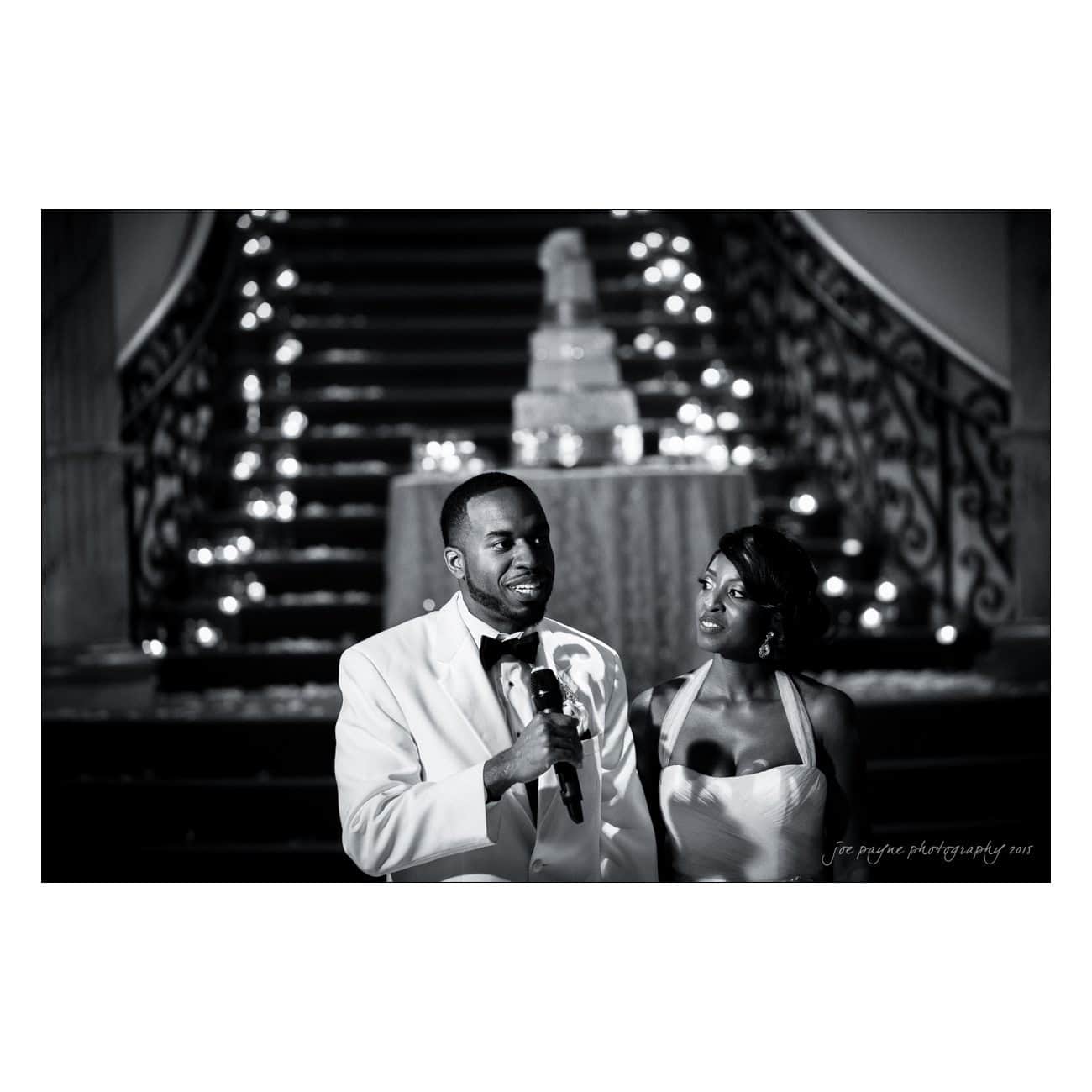 A couple in formal attire stands in front of a grand staircase with a tiered cake in the background. The man holds a microphone, and they appear to be at a wedding reception. String lights adorn the staircase. The image is in black and white.