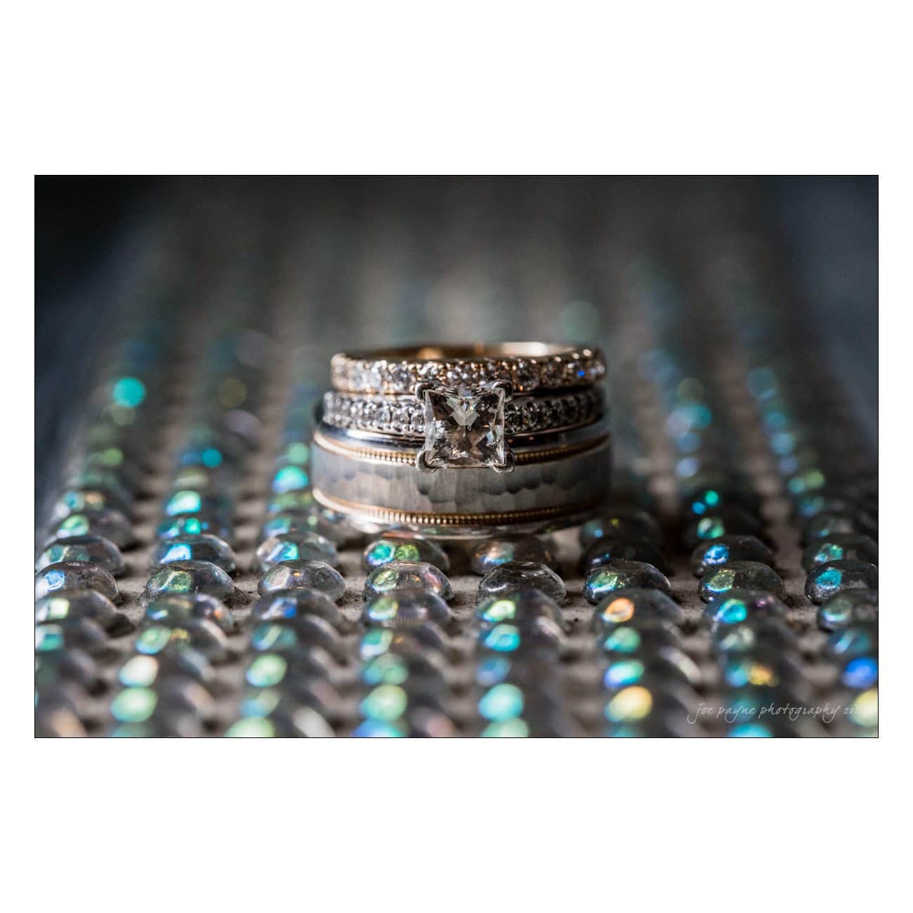 A stack of elegant rings, including a diamond engagement ring, displayed on a reflective, colorful beaded surface. The rings feature intricate metalwork and shimmering stones, creating a luxurious and vibrant appearance.