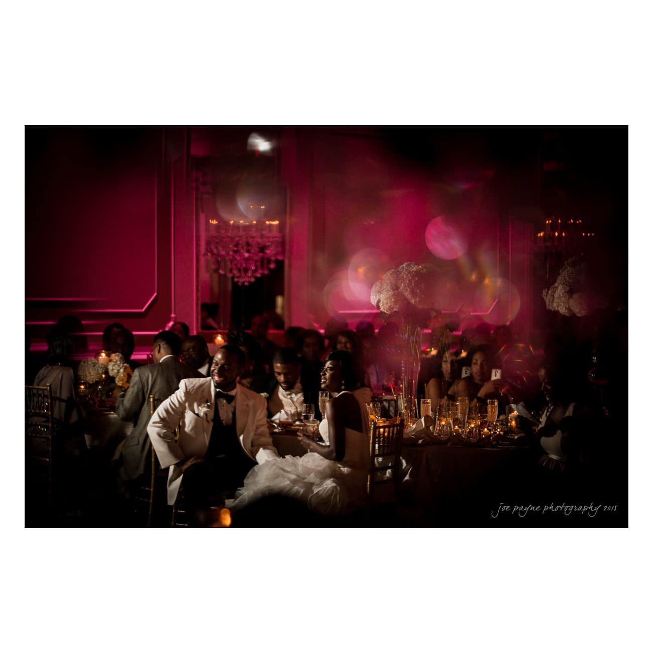 A lavish wedding reception with guests seated at tables adorned with elegant centerpieces. The room is dimly lit, casting a pinkish glow. A couple in wedding attire is seated prominently, surrounded by guests enjoying the celebration.