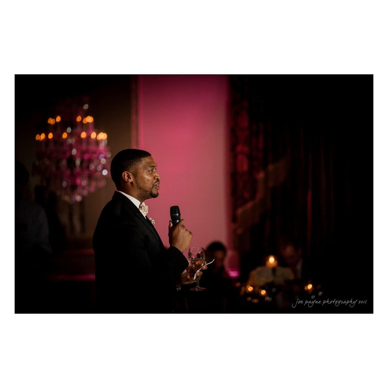 A person in formal attire holds a microphone and a drink, addressing an audience in a dimly lit room with elegant chandeliers and a pinkish glow. The setting suggests a formal event or celebration.