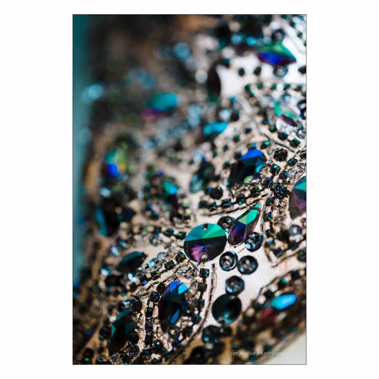 Close-up of an ornate fabric adorned with multicolored, iridescent gemstones and intricate embroidery. The gems reflect shades of blue, green, and purple, creating a luxurious and detailed texture.