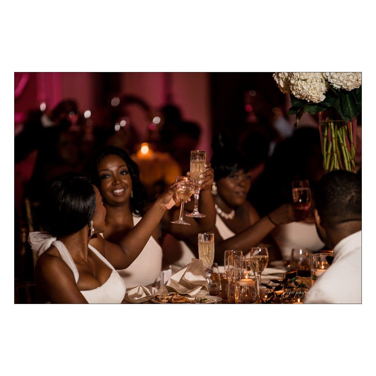 A group of people in formal attire raise their champagne glasses in a toast during a celebration. The table is elegantly set with candles and decorations, and the background is softly lit, creating a warm and festive atmosphere.