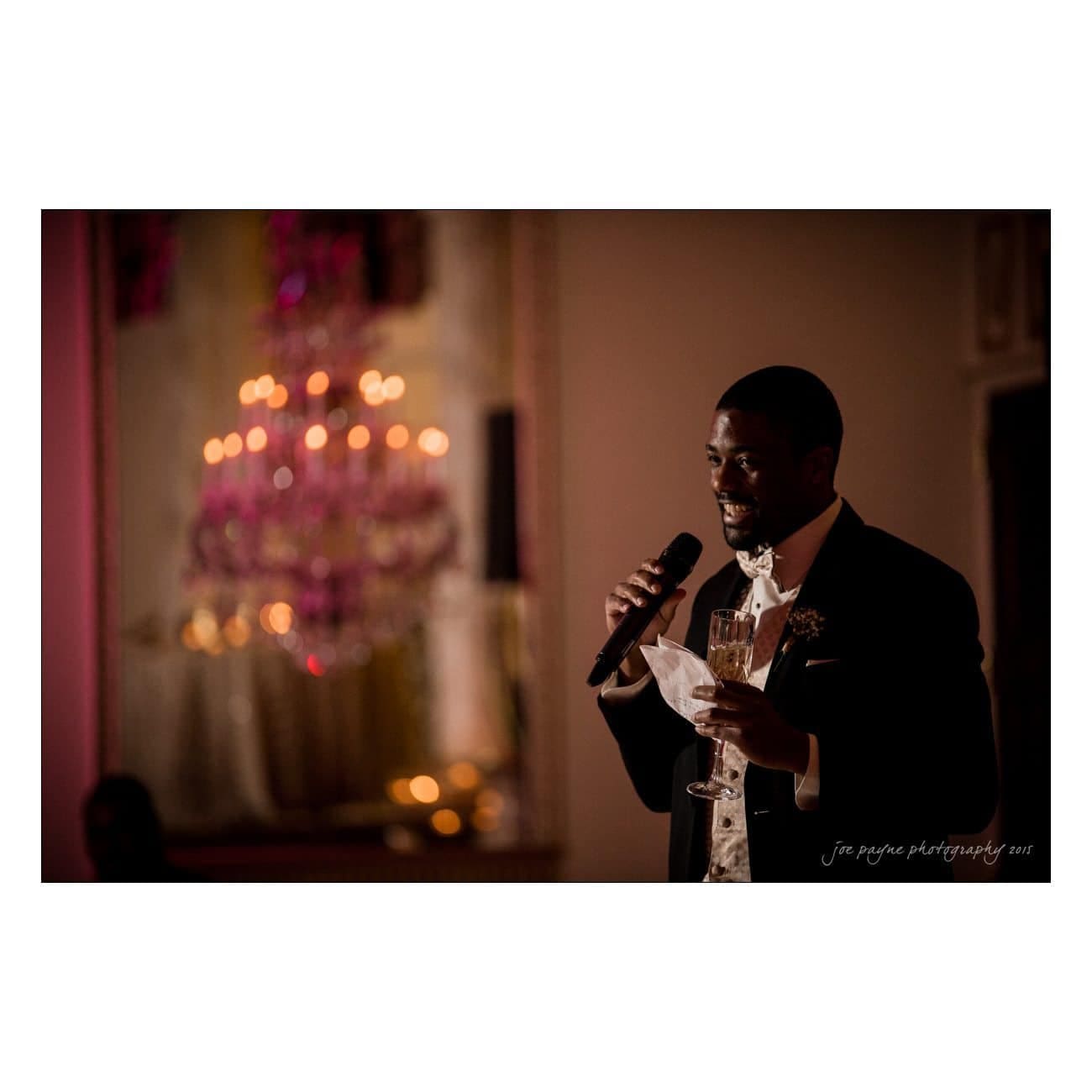 A person in a formal black suit with a bow tie holds a microphone and a champagne glass while speaking. A softly lit, elegant chandelier is blurred in the background, adding a warm atmosphere to the scene.