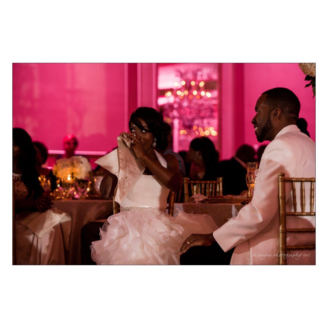 A bride wipes her tears while sitting next to the groom at a wedding reception. The room is decorated with pink lighting and elegant table settings. Guests are seated and watching the couple.