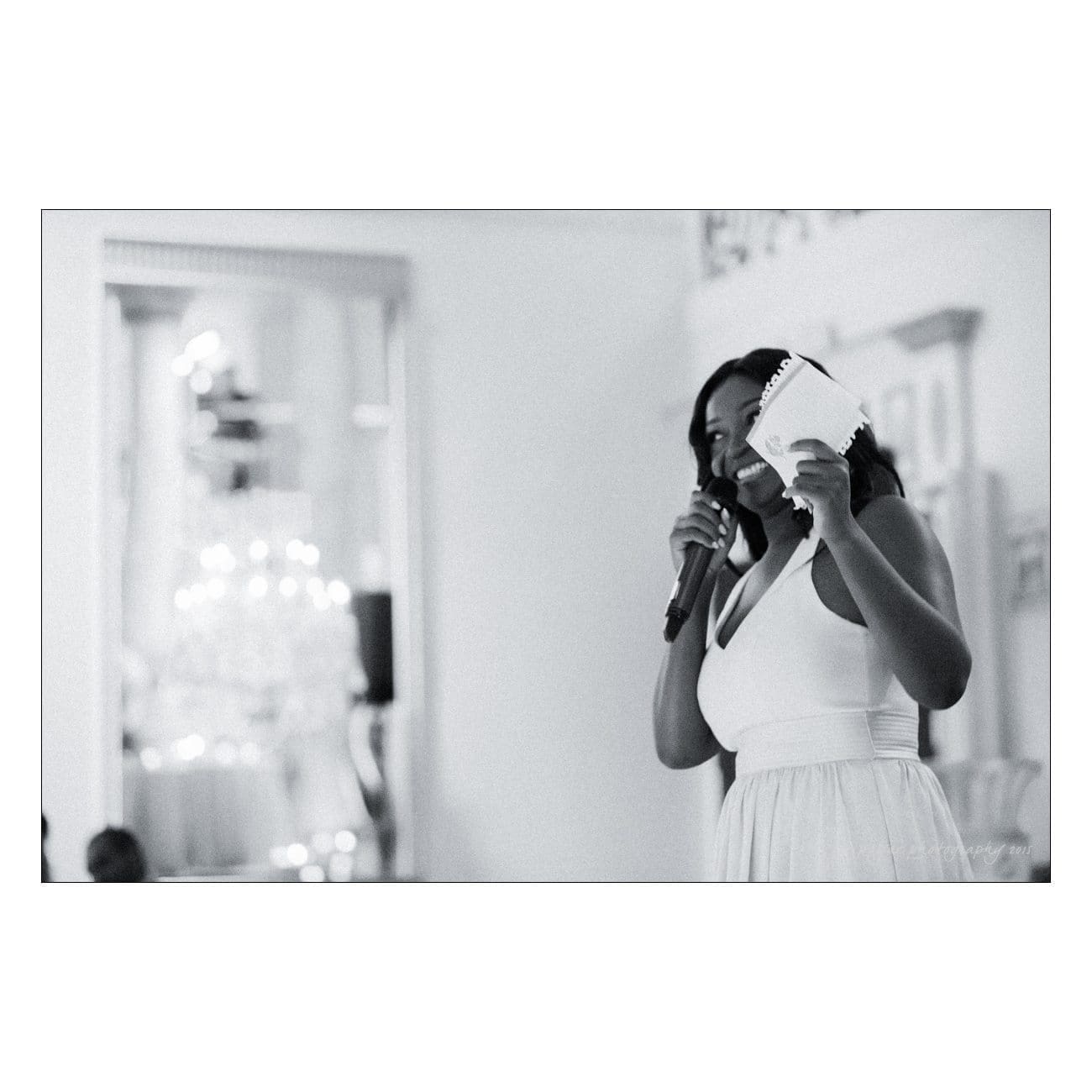 A woman in a white dress speaks into a microphone, holding a piece of paper, in a softly lit room. A chandelier is visible in the background. The image is in black and white.