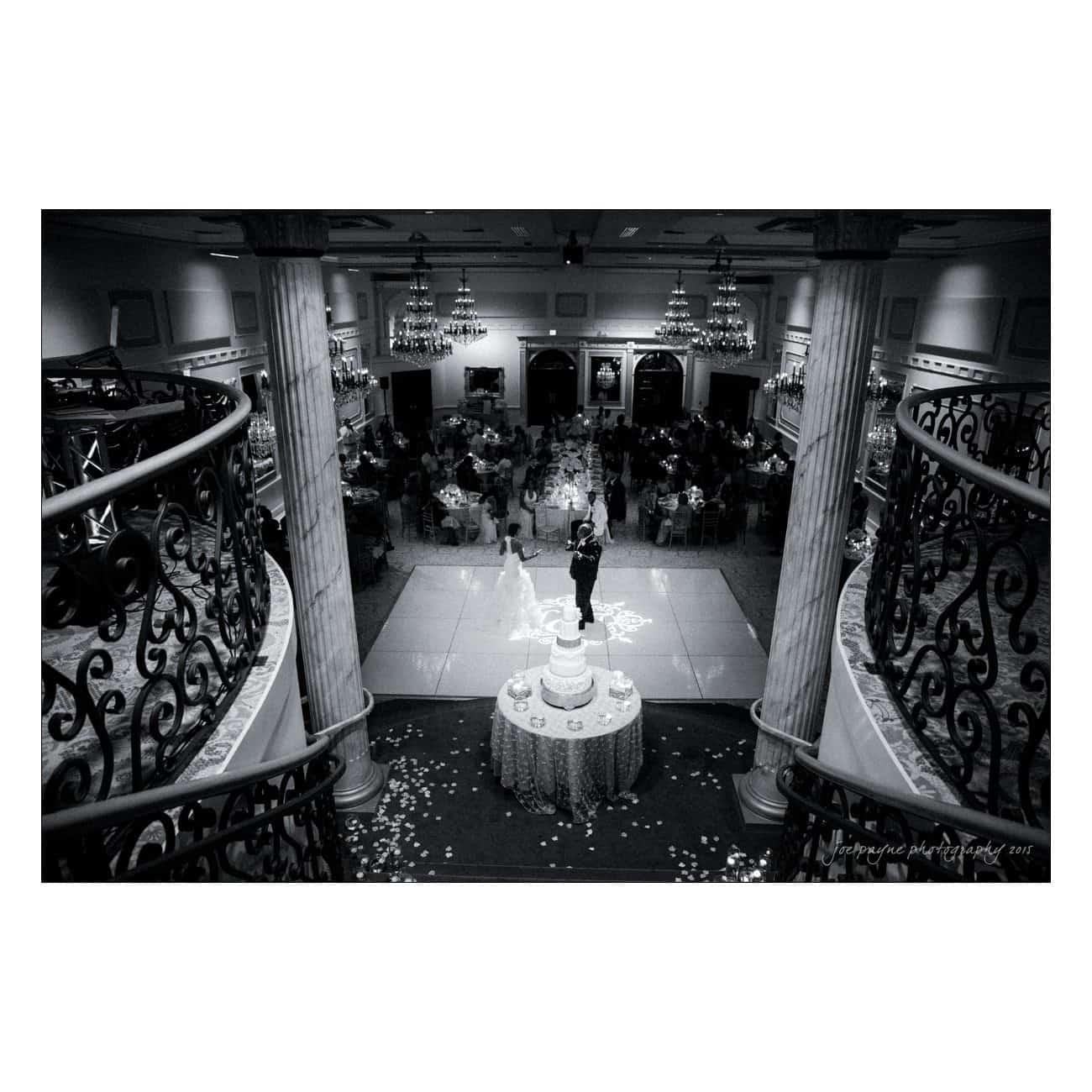 A black and white image of a wedding reception. A couple dances on a square dance floor, surrounded by seated guests. A tiered cake is prominently displayed in the foreground, and elegant chandeliers hang from the ceiling.