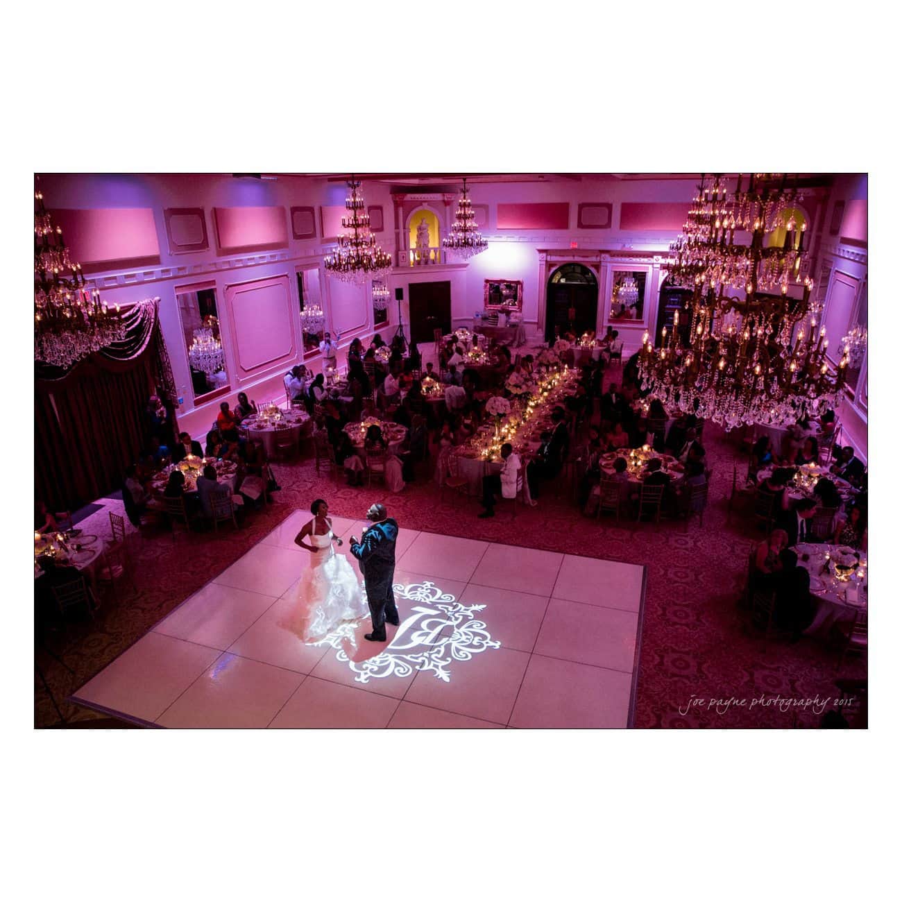 A bride and groom dance on a monogrammed floor in an elegant ballroom, illuminated by soft lighting. Guests sit at round tables adorned with candles and floral arrangements. Crystal chandeliers add a luxurious touch to the scene.