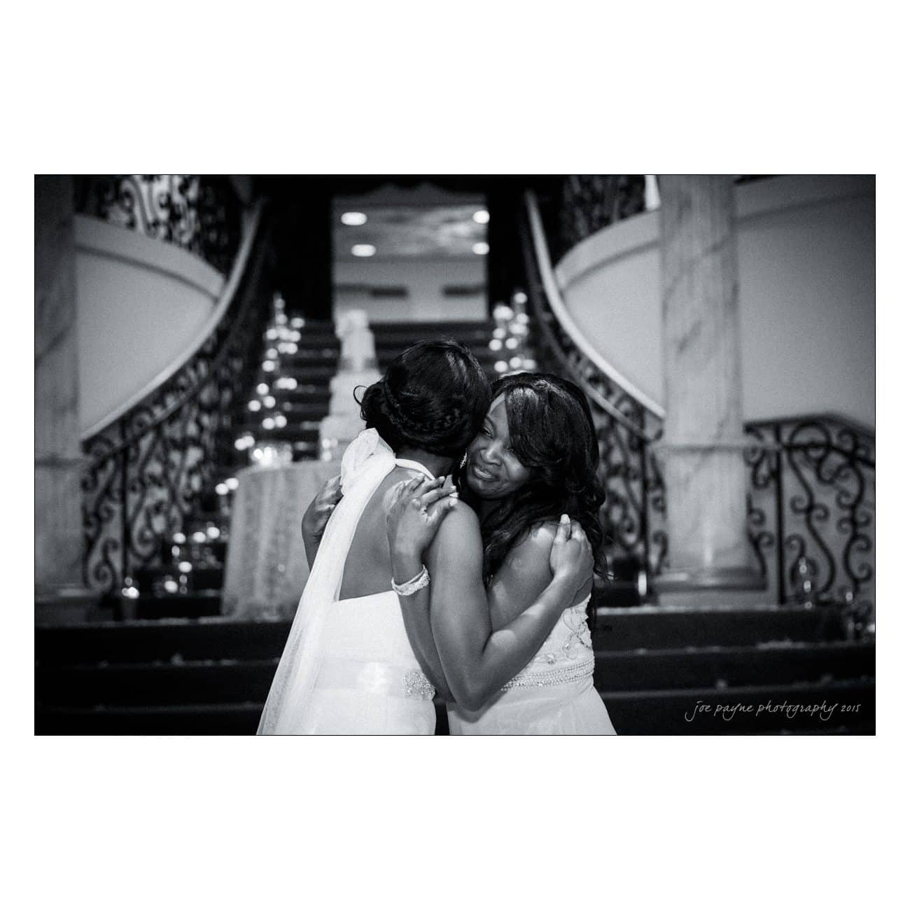 Two women embrace on an ornate staircase lined with candles. Both wear elegant dresses, and the background features intricate railings and marble columns. The atmosphere is warm and intimate.