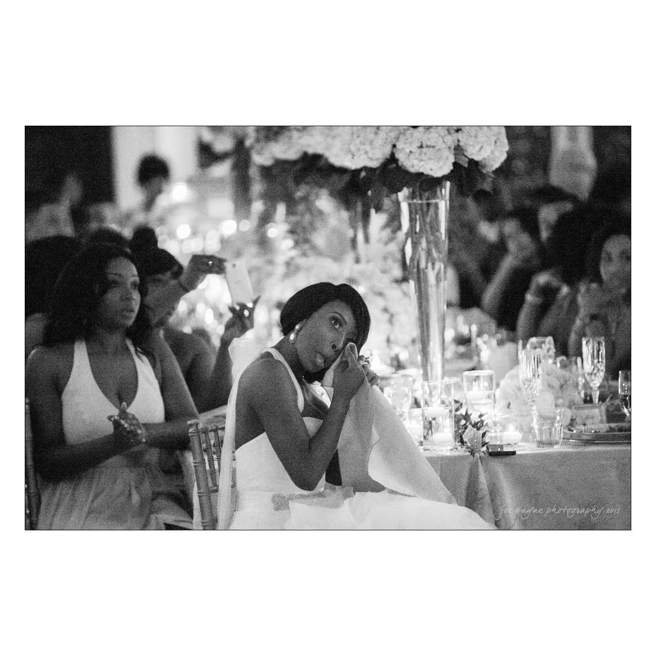 A black and white photo of a woman in a formal dress looking emotional and wiping her eye with a napkin. She is seated at an event with elegantly set tables and a large floral arrangement nearby. Other attendees are visible in the background.