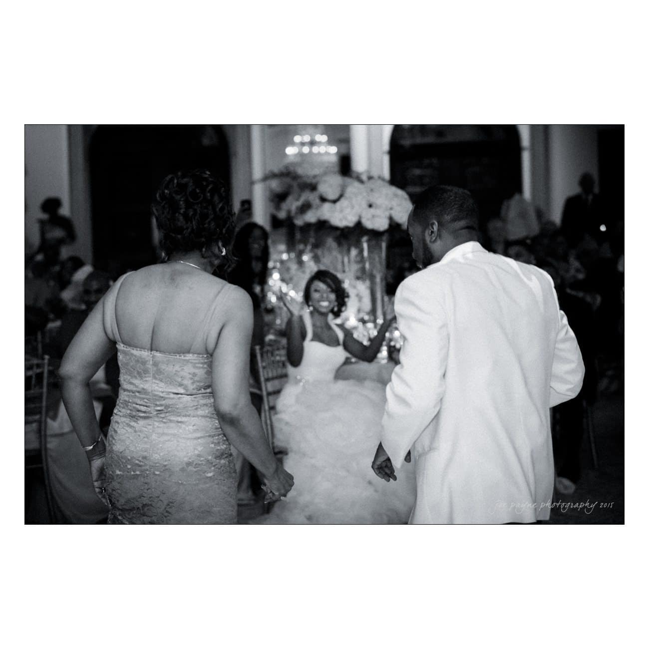 A black and white photo of a bride sitting and smiling as two people, dressed in formal attire, approach her in a dimly lit reception hall. There are guests and floral decorations in the background. The atmosphere is festive and elegant.