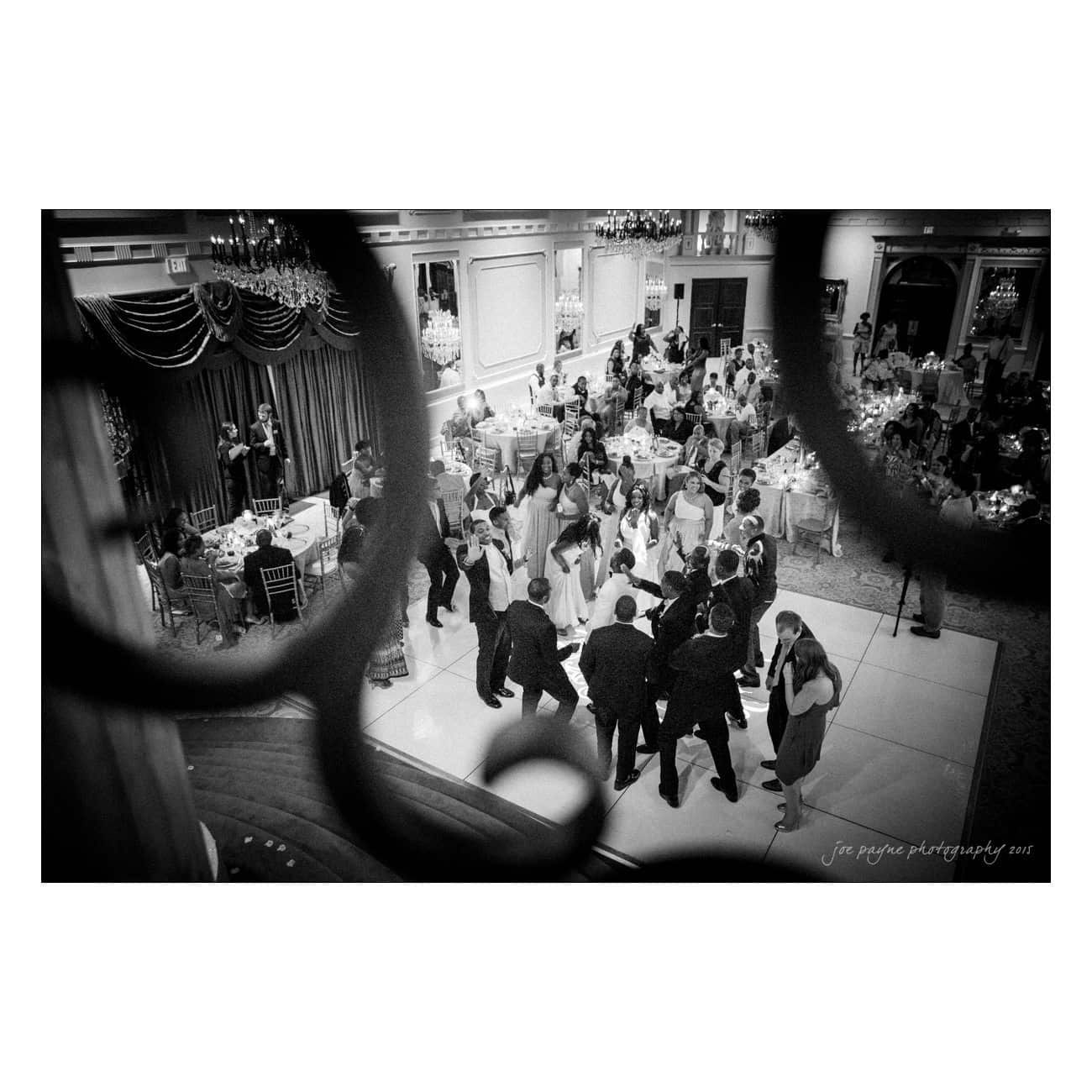 A black and white photo of a lively wedding reception. People are dancing in the center of the image, surrounded by elegantly set tables. The scene is viewed through decorative circular railings in the foreground.