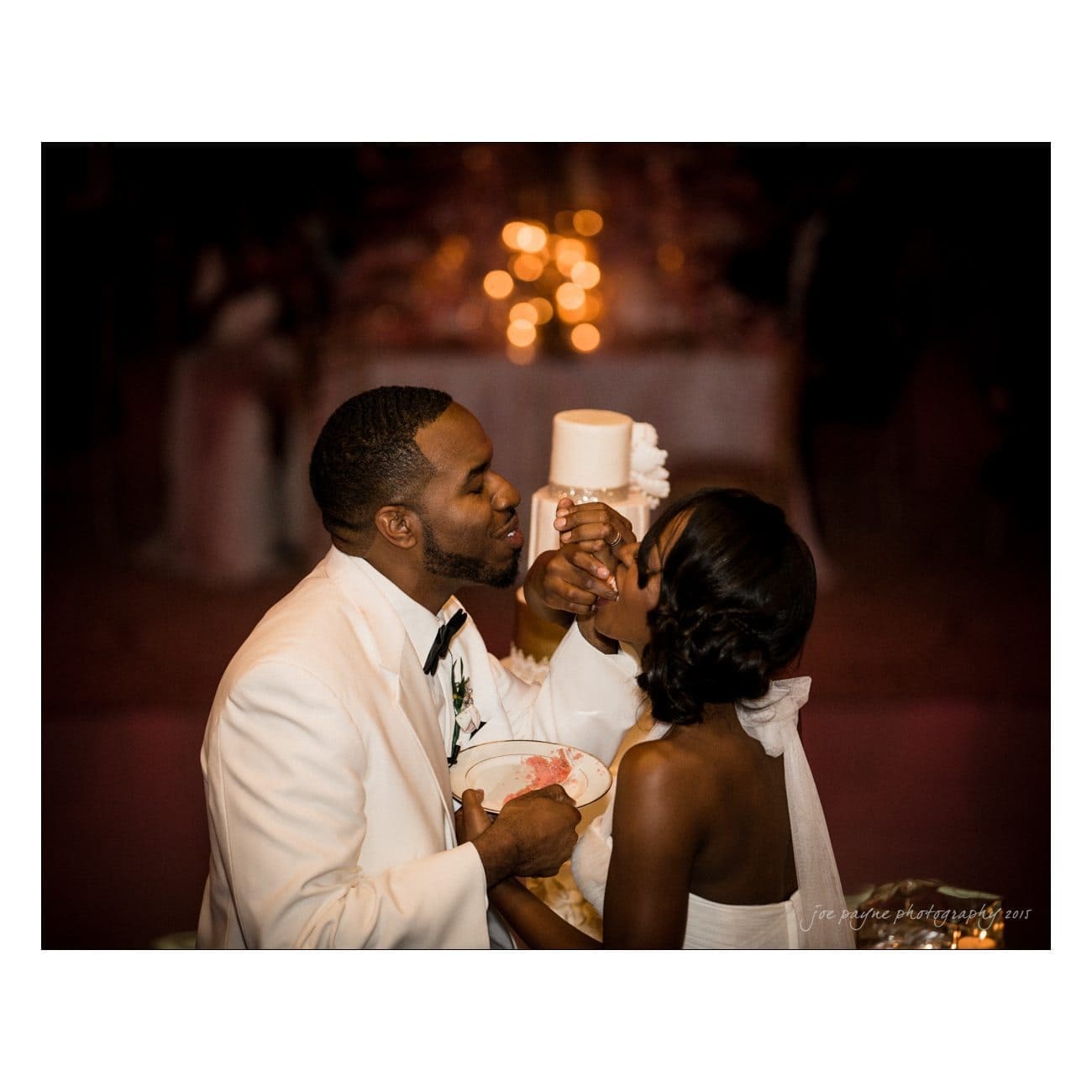 A bride and groom in formal attire share cake, feeding each other tenderly. The groom is in a white tuxedo, and the bride is in a wedding dress. They are surrounded by a softly lit, romantic setting.