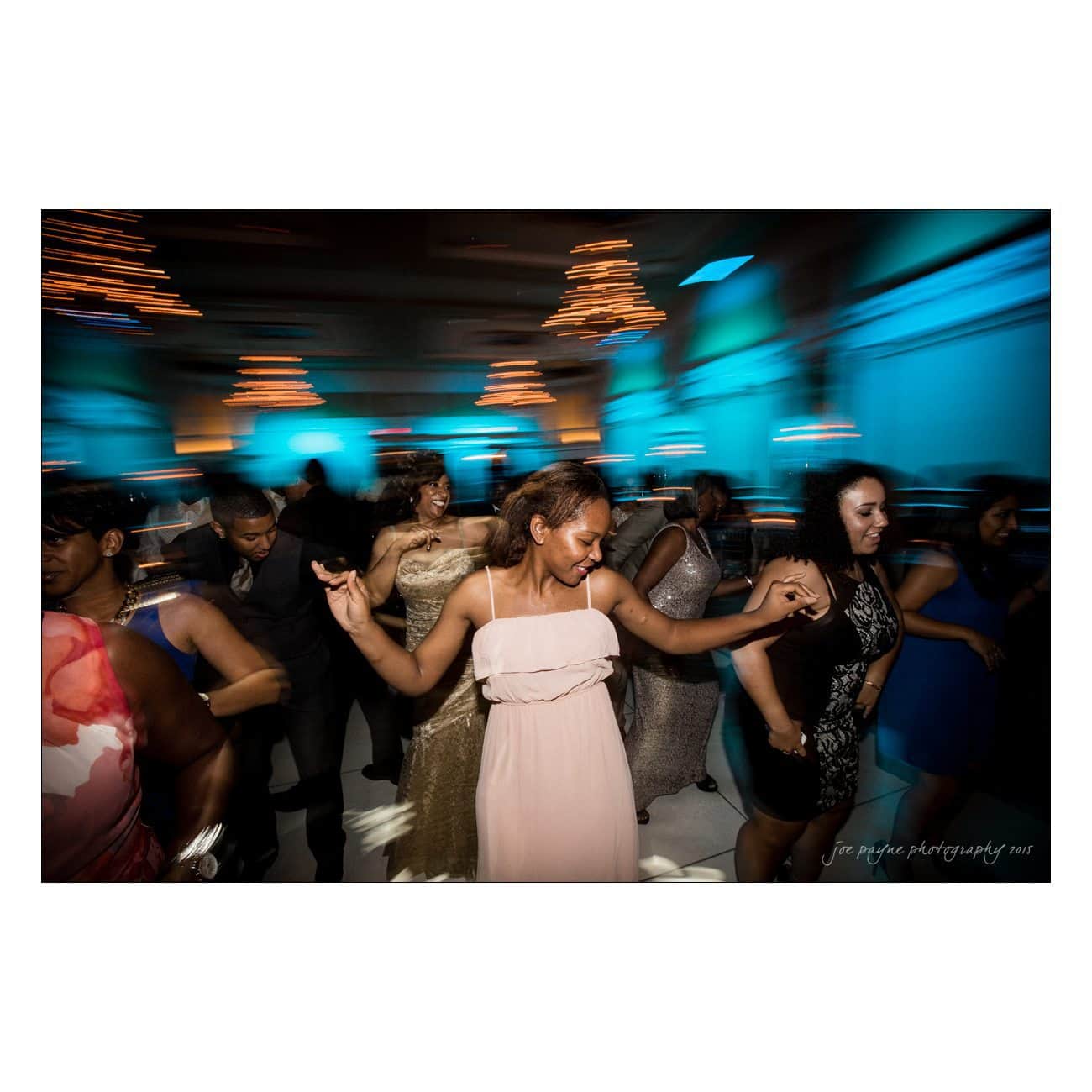A group of people are dancing joyfully at an indoor event. The background features glowing blue and warm lights, creating a lively atmosphere. Most are wearing formal attire, with one woman in a light pink dress smiling at the center.