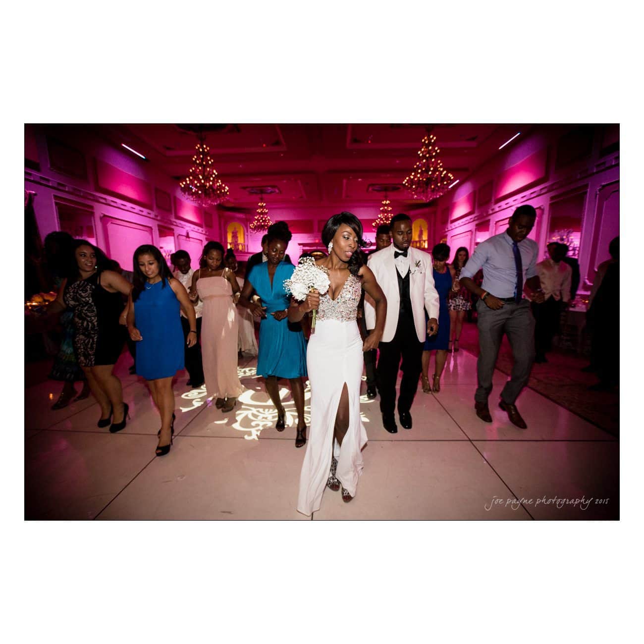 A bride in a white dress, holding a bouquet, dances joyfully on the dance floor with guests. The venue is dimly lit with pink lighting, creating a festive atmosphere. People around her are smiling and enjoying the celebration.