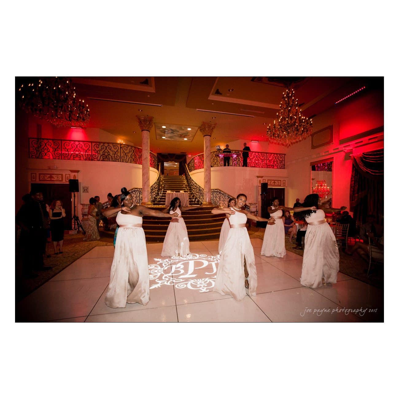 A group of young girls in elegant white dresses performs a dance on a decorated ballroom floor, surrounded by ornate architecture and warm red lighting. A grand staircase is visible in the background.