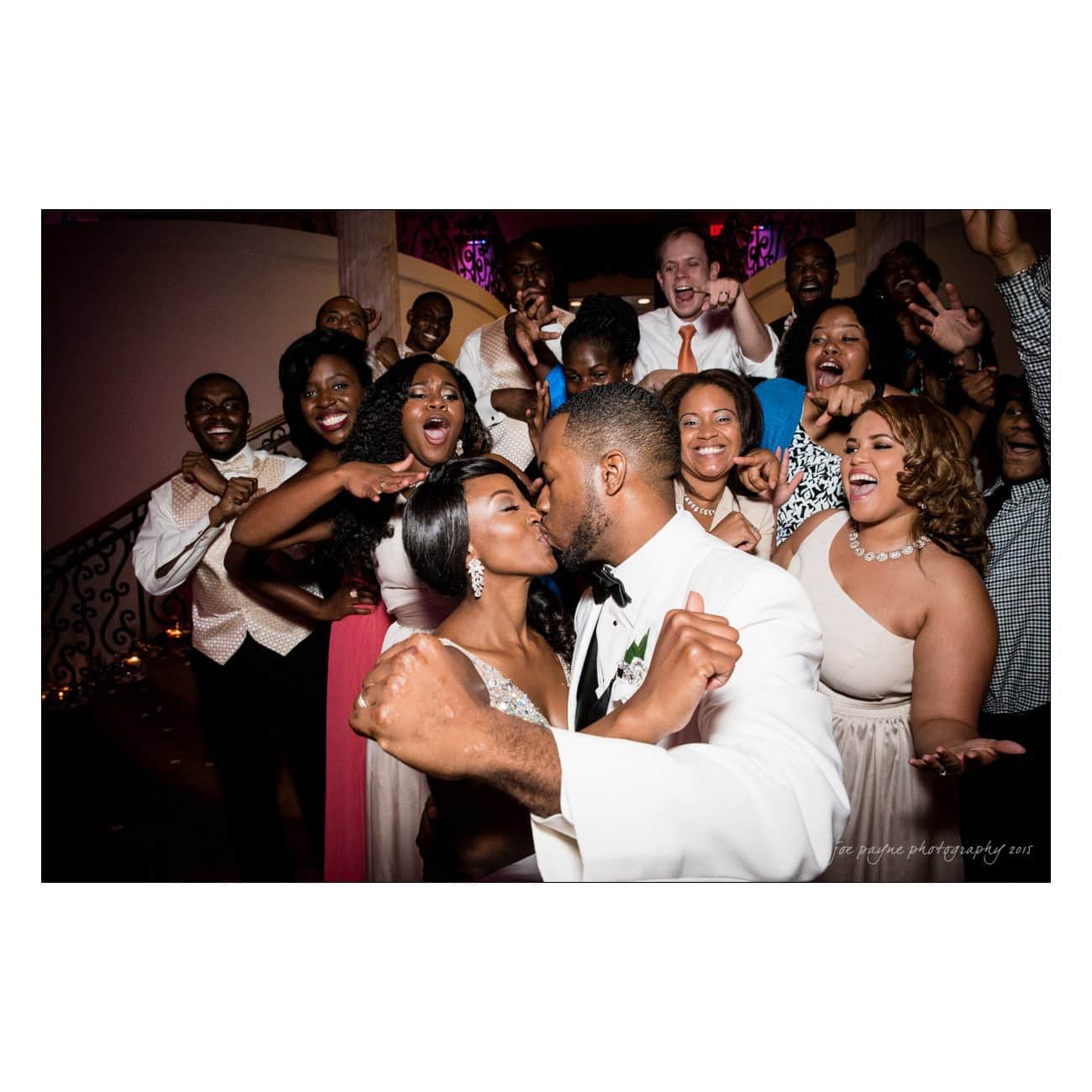A newlywed couple shares a kiss surrounded by cheering friends and family. Theyre dressed in wedding attire, and everyone appears joyful and celebratory, with arms raised in excitement.